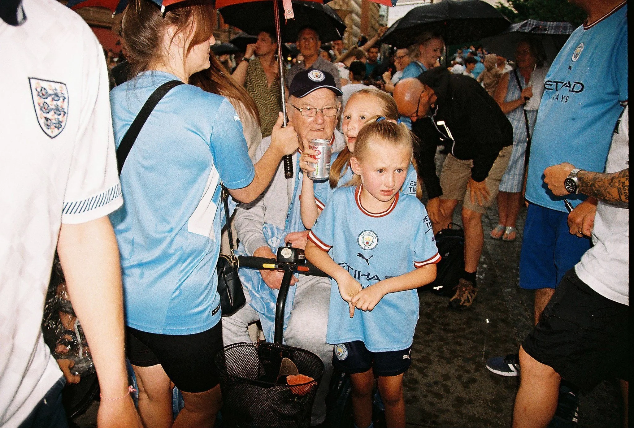 Group of Manchester City football fans, including children, gathered outdoors with umbrellas, wearing light blue shirts, during a rainy day.