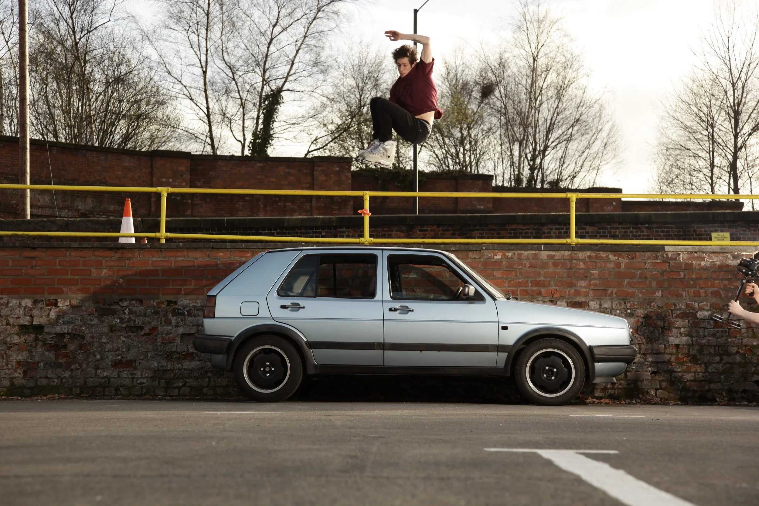 A person jumping off a yellow railing above a silver car parked against a brick wall.