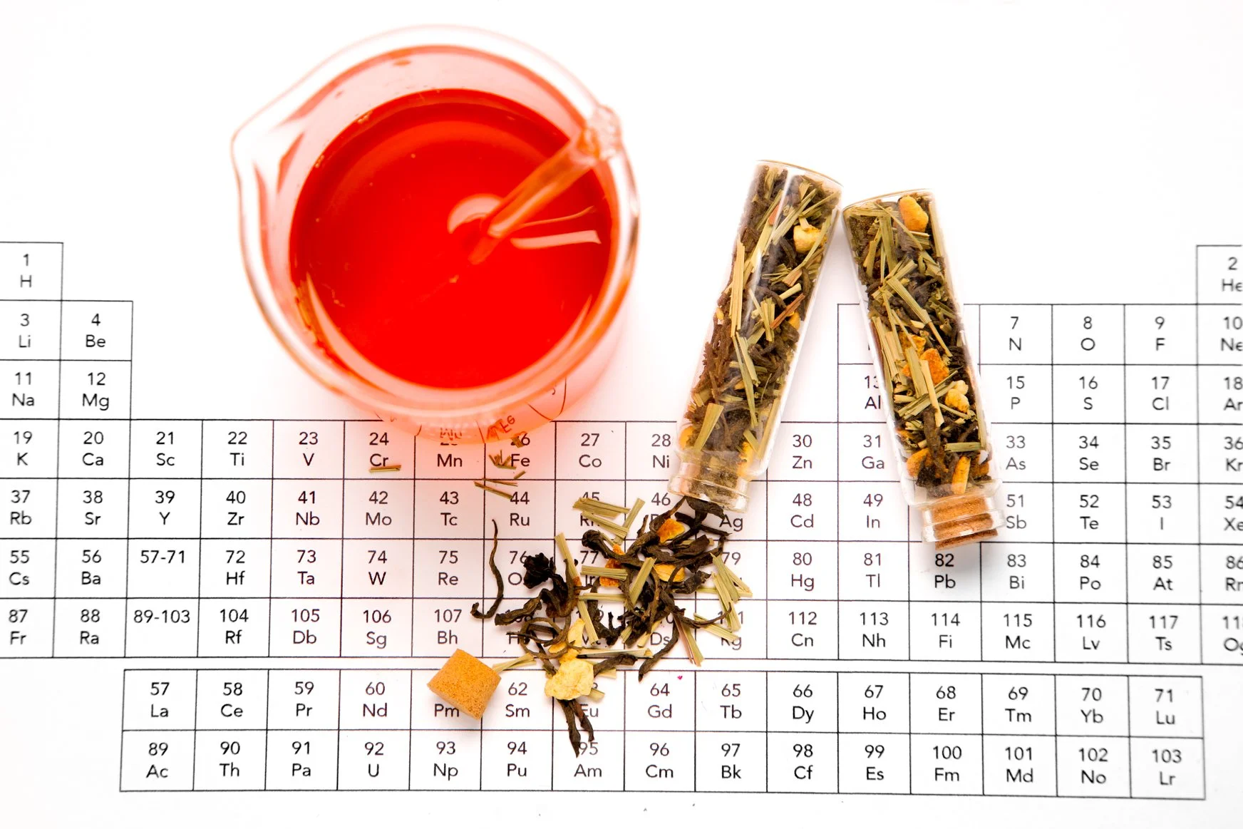 A glass of red herbal tea, two containers of loose herbal tea, and scattered loose tea leaves and a tea bag on top of a periodic table of elements.