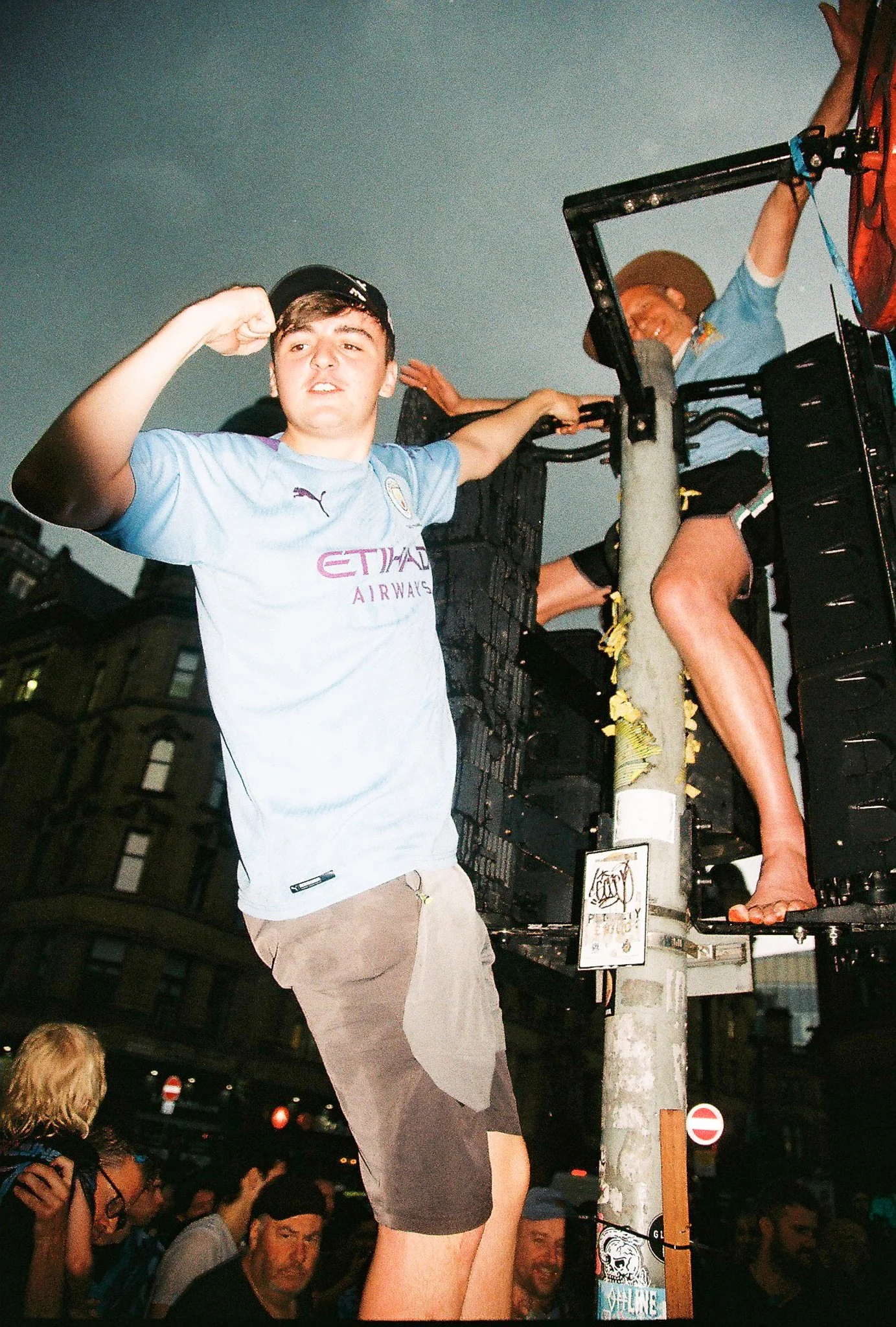 A young man in a light blue soccer jersey is standing with one arm raised in a fist, surrounded by a crowd on the street at night. He is wearing shorts and a baseball cap. There is a woman sitting on a platform or structure behind him, with her legs 