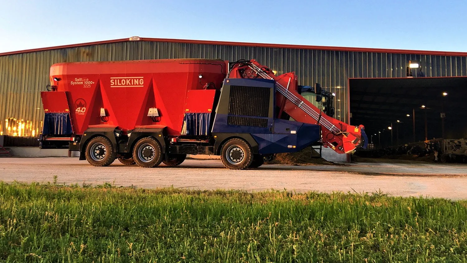 Side profile view of a massive 3-axle Siloking SelfLine System 1000 plus 3225 self-propelled triple-auger mixer wagon parked outside a large illuminated commercial dairy barn at dusk.