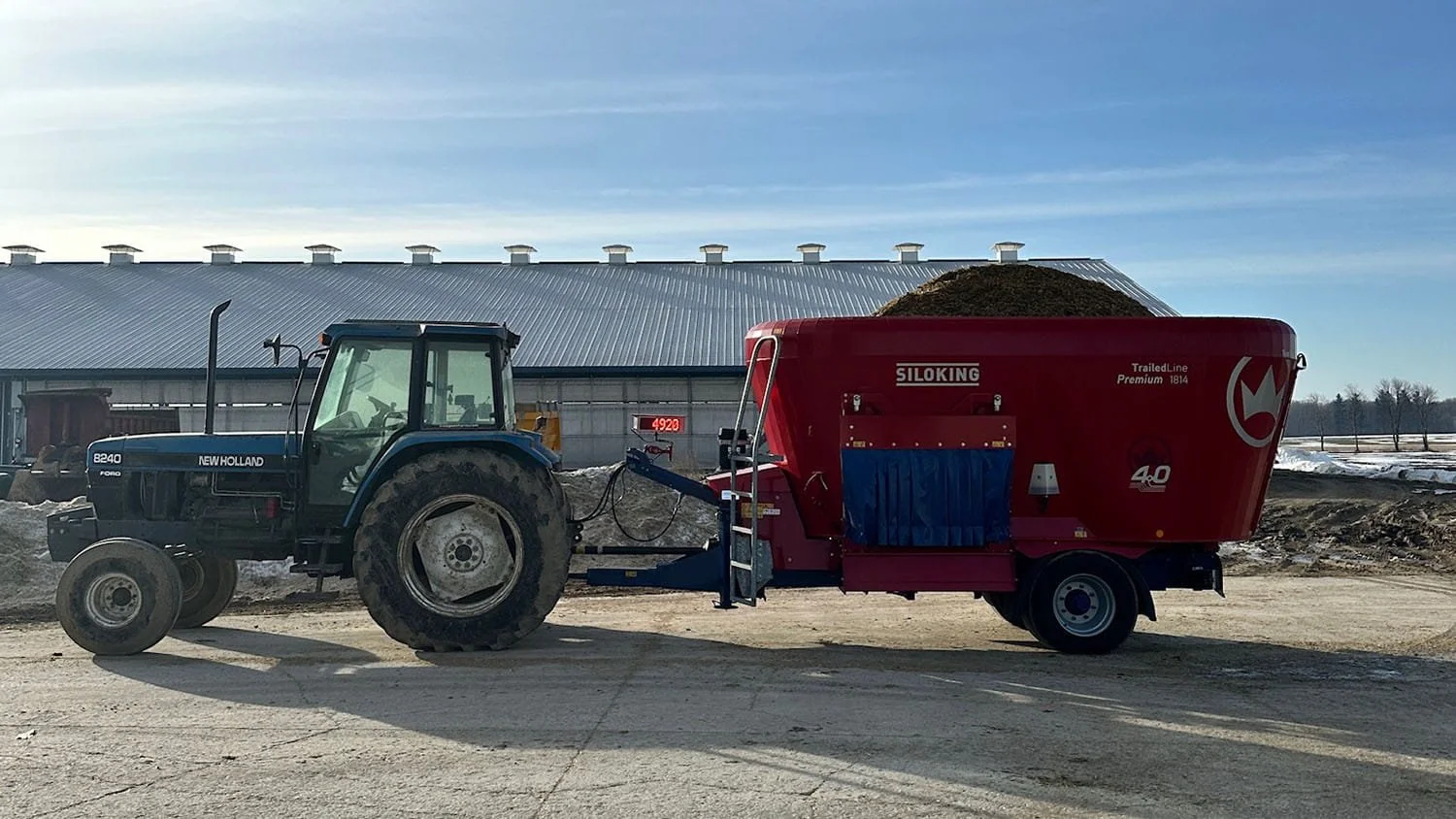 A red Siloking TrailedLine Premium 1814 vertical mixer wagon hitched to a blue Ford New Holland tractor in a farmyard.