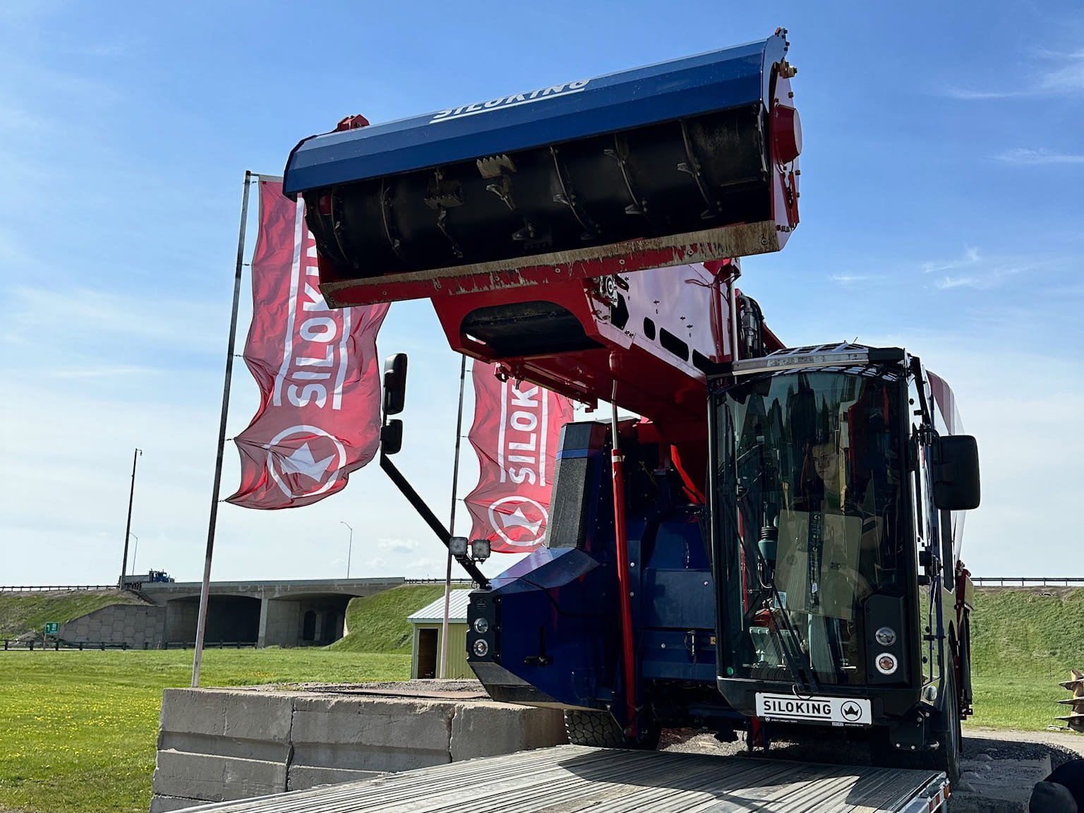 Front view of an operator inside a blue Siloking self-propelled vertical mixer cab with the loading arm raised high next to red corporate flags.