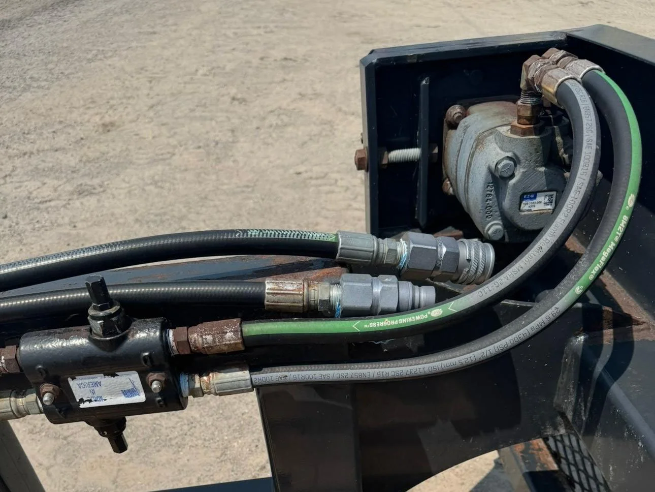 Detailed close-up view of the hydraulic drive motor, flow control valve, and quick-disconnect line couplers on a skid steer bunk shaver attachment.