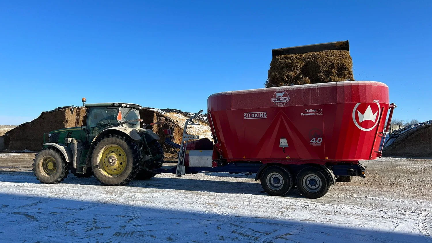 A green John Deere tractor loading silage into a 26 cubic meter Siloking TrailedLine Premium 3022 vertical mixer wagon on a snow-covered farm lot in winter.