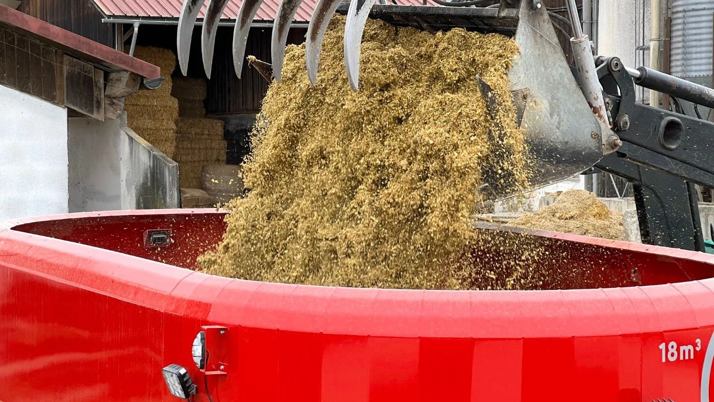 A farm front loader dumping fresh silage into the open mixing tub of an 18m³ electric Siloking TruckLine eTruck 2012 vertical mixer wagon.