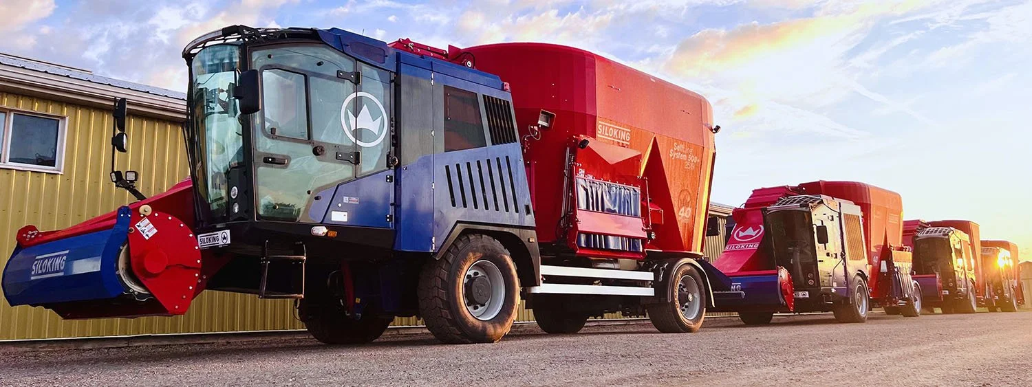 A long line of red and blue Siloking SelfLine System 500 self-propelled mixer wagons parked alongside a dealership service center workshop at sunset.