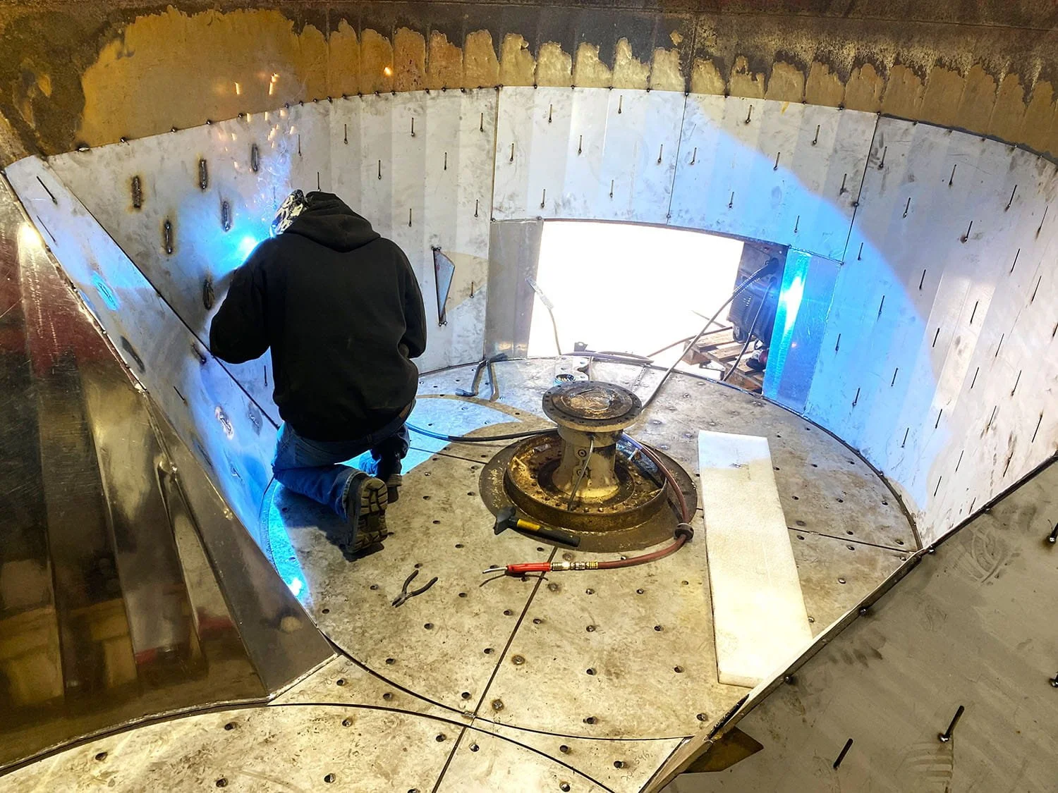 A welder in a protective mask performing precision structural welding to install stainless steel wear plates inside a vertical mixer tub.