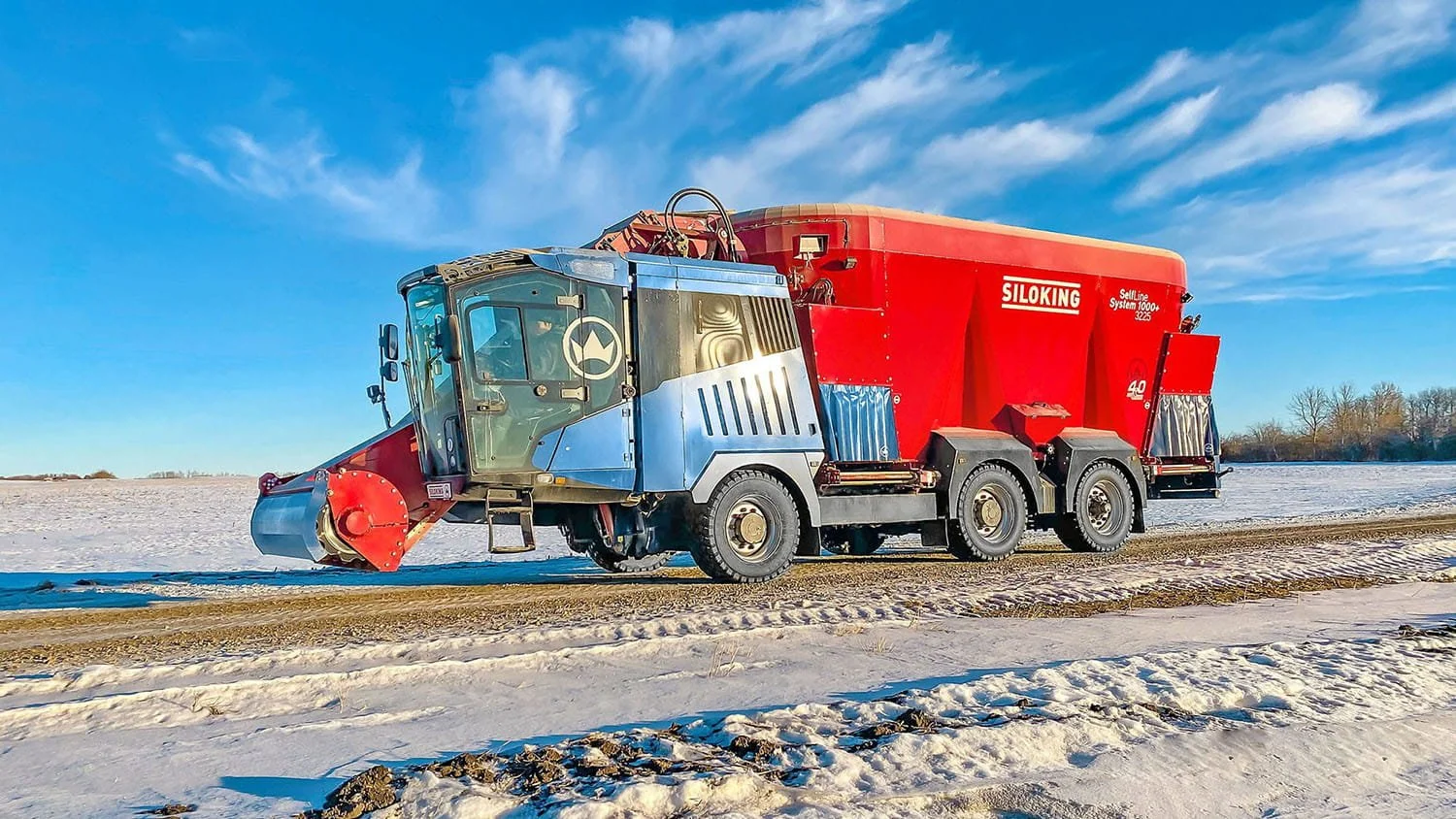 A red and blue Siloking SelfLine System 1000 plus triple-axle TMR mixer travelling smoothly down a snow-packed farm lane under a clear blue sky.