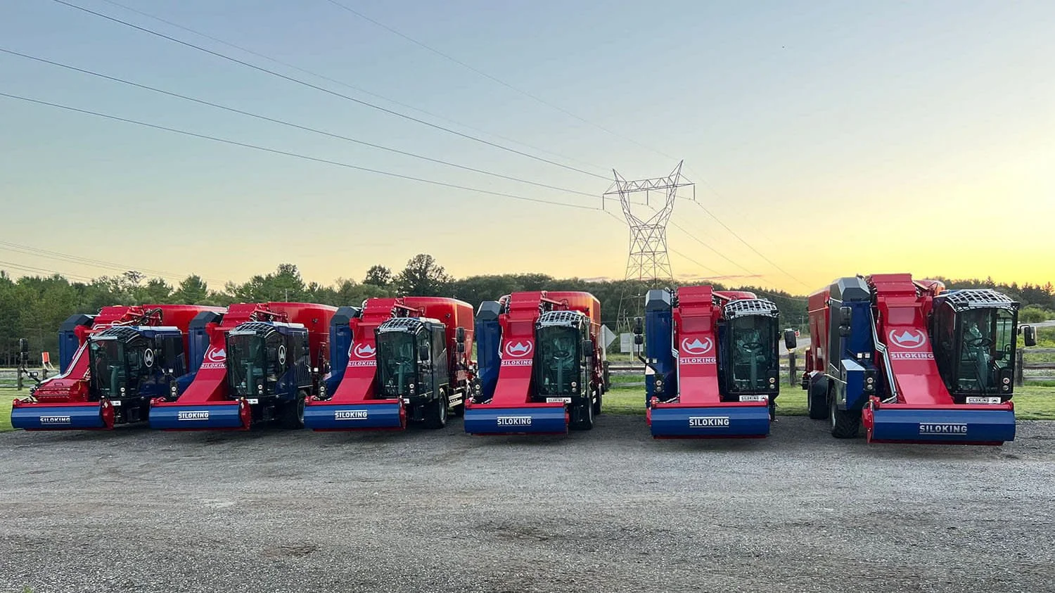 A lineup of red and blue self-propelled vertical feed mixers parked at the Siloking Canada headquarters during sunset.
