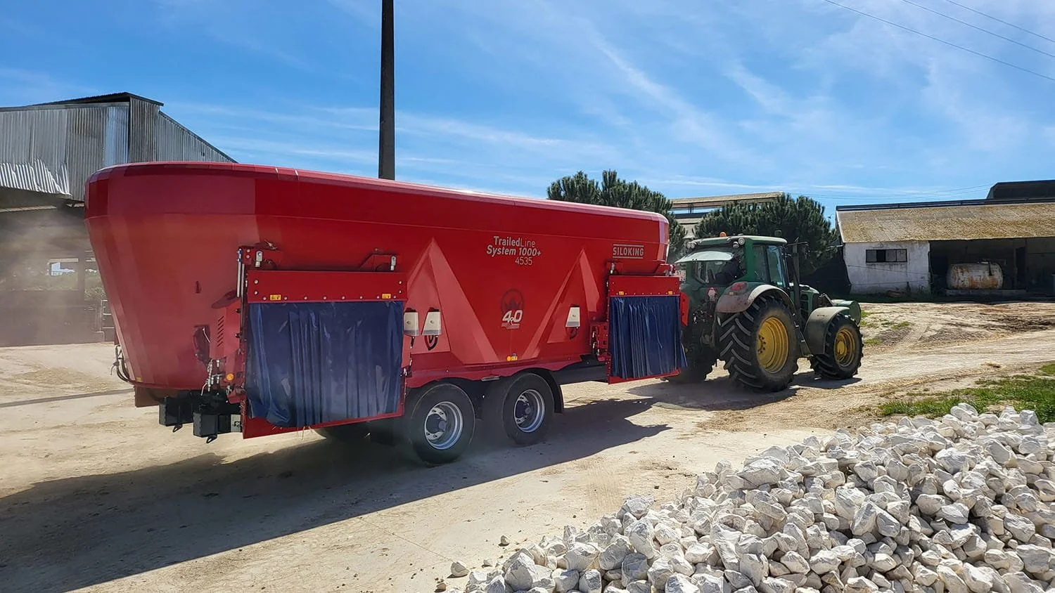 A green John Deere tractor towing a massive Siloking TrailedLine System 1000 plus 4535 pull-type feed mixer past a farm building on a sunny day.