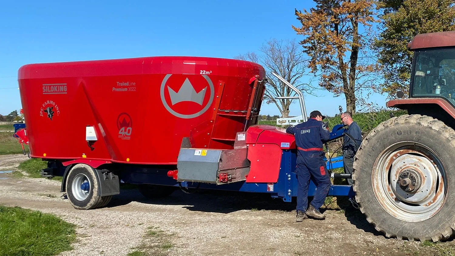 An agricultural service mechanic and the farmer performing diagnostic repairs on the hitch and chassis of a red Siloking TrailedLine Premium 3022 mixer wagon out in a farm field yard.