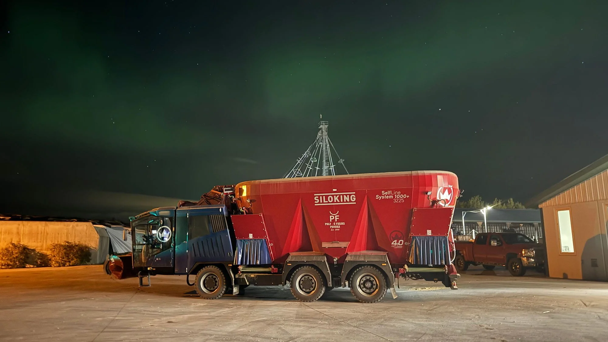 A massive Siloking SelfLine System 1000 plus 3225 vertical feed mixer at a dairy farm under a night sky lit by green aurora borealis.

