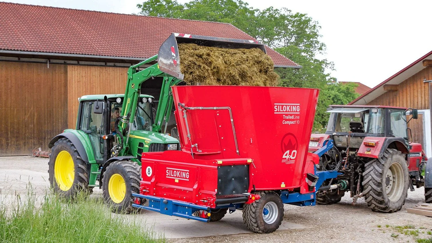 A red Siloking TrailedLine Compact 10 mixer wagon being loaded with silage by a green John Deere farm tractor with a front-loader bucket in a farmyard.