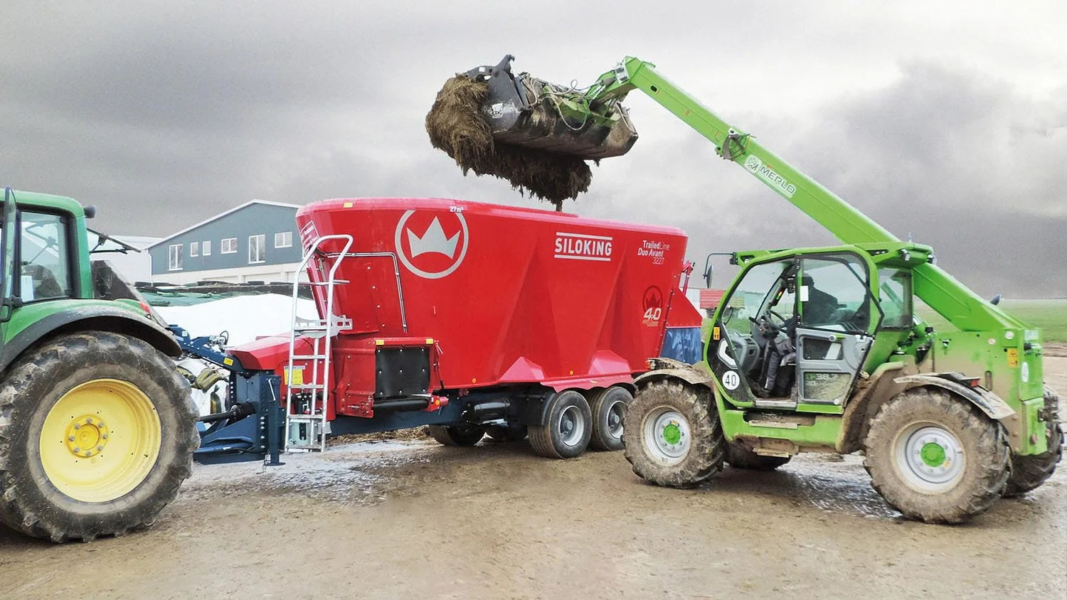 A green Merlo telehandler loading dense silage into a massive red Siloking TrailedLine Duo Avant 3227 twin-auger mixer wagon next to a John Deere tractor in a muddy winter yard.