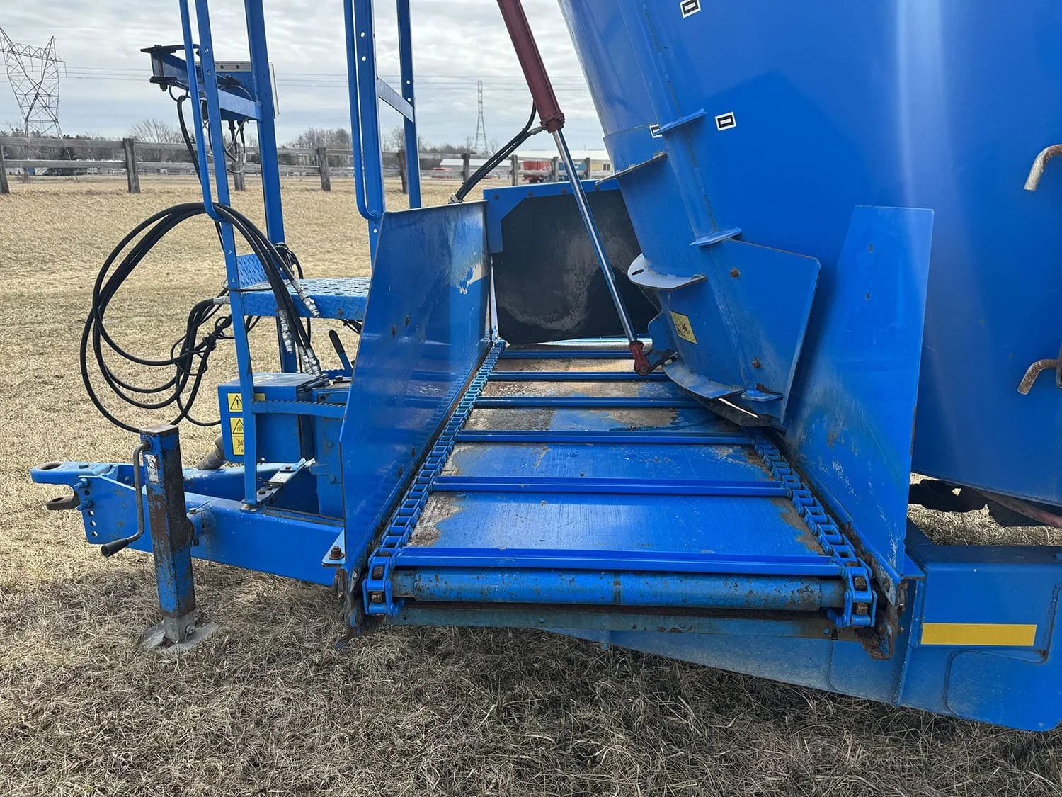 Close-up of the front cross conveyor discharge chain and hydraulic lines on a pull type Patz 2400 Series II 650XH cattle feed mixer.