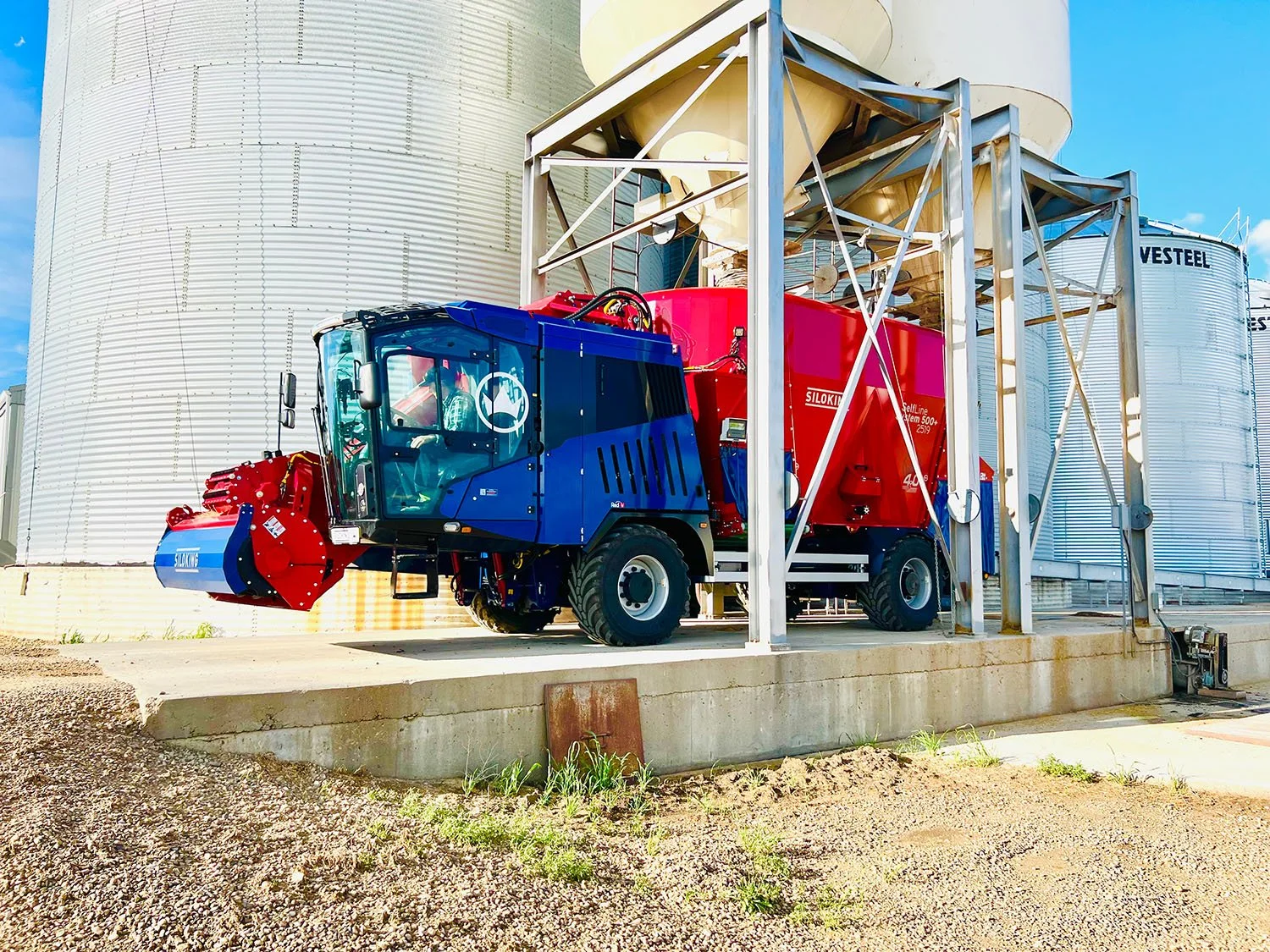 A red and blue Siloking SelfLine System 500+ 2519 self-propelled vertical TMR feed mixer parked on a concrete pad underneath an overhead grain bin hopper on a farm.