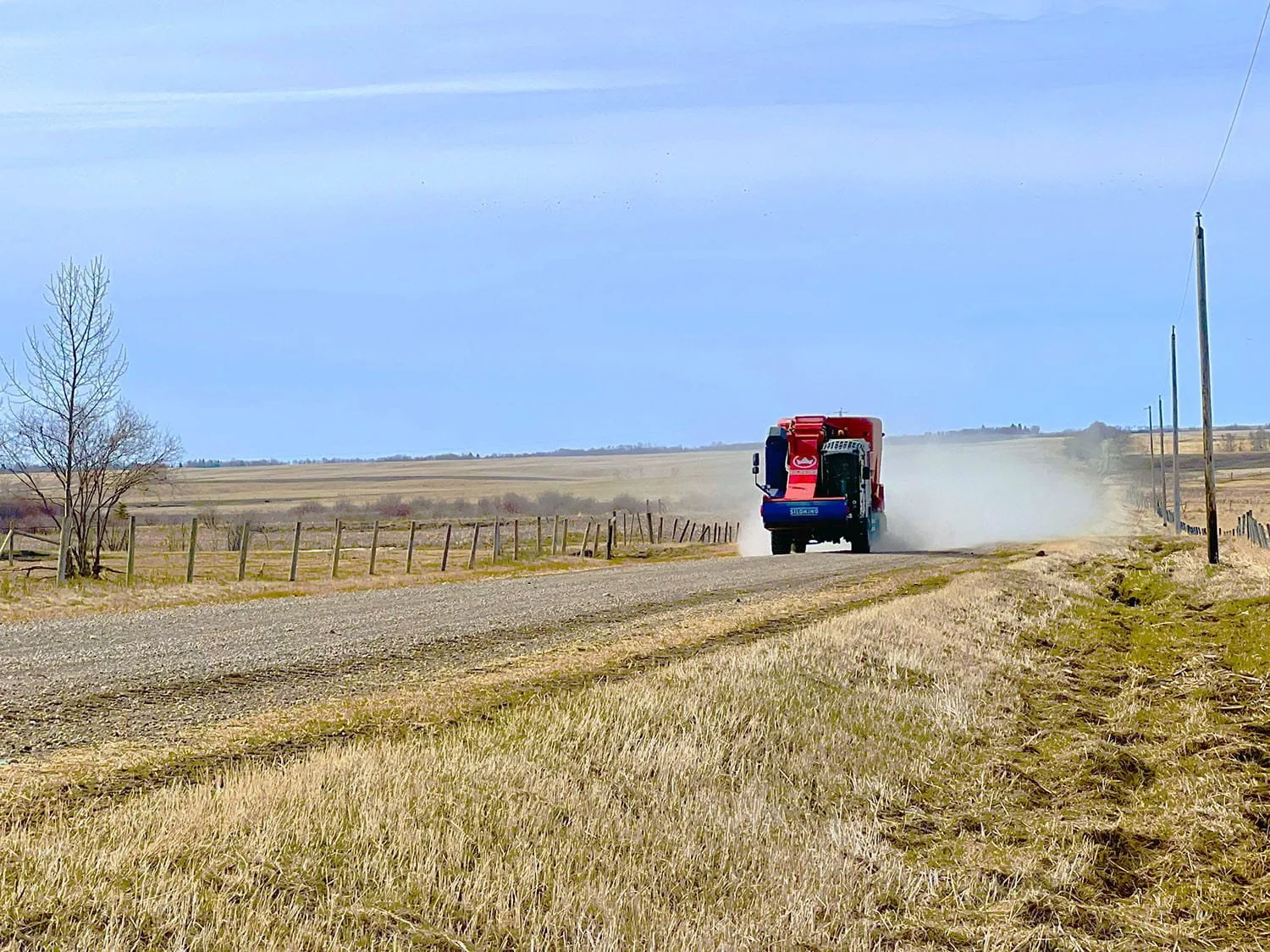 A red and blue Siloking SelfLine System 500+ self-propelled vertical TMR feed mixer driving down a gravel road through open farm fields, kicking up a dust cloud behind it.