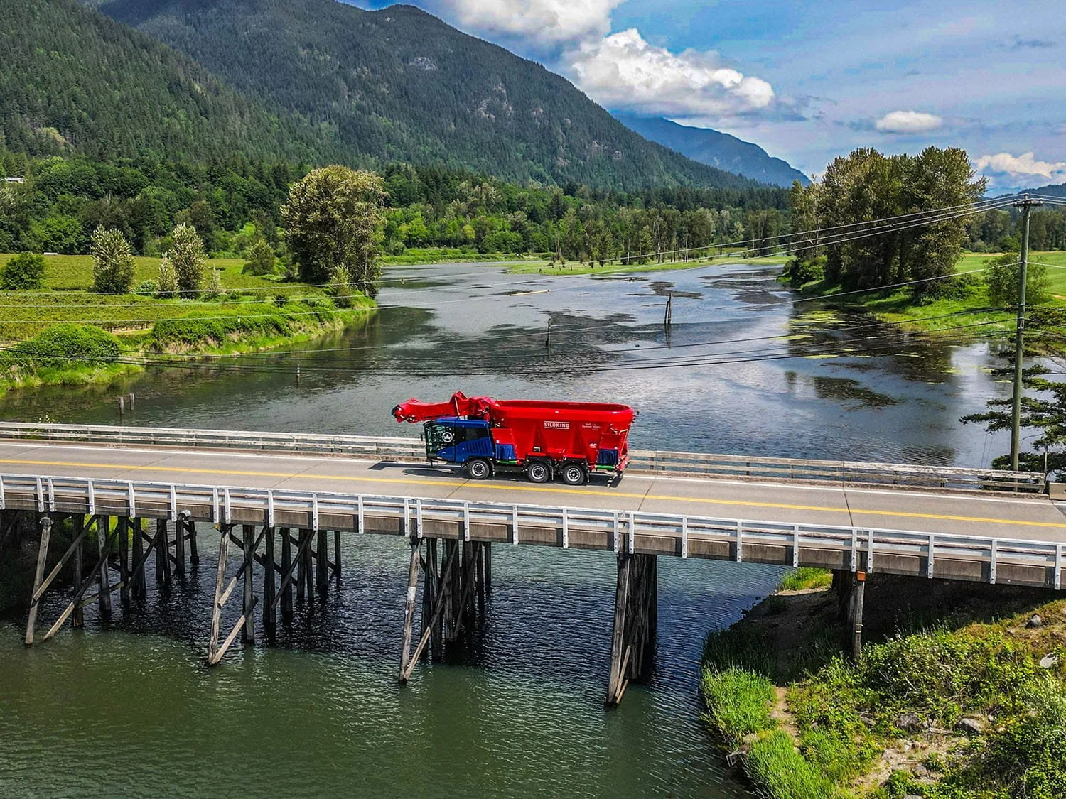 An aerial landscape view of a Siloking SelfLine self-propelled feed mixer driving across a bridge in a rural agricultural region.