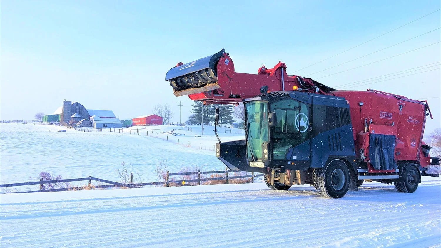 A heavy-duty red and blue Siloking SelfLine System 500 plus driving down a snowy rural concession route past traditional wooden barn structures.