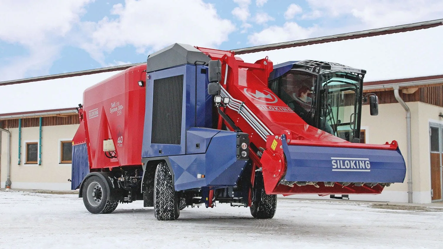 Front angle profile view of a red and blue Siloking SelfLine System 300 plus 2519 vertical feed mixer operating on a snow-cleared farm lot in winter.