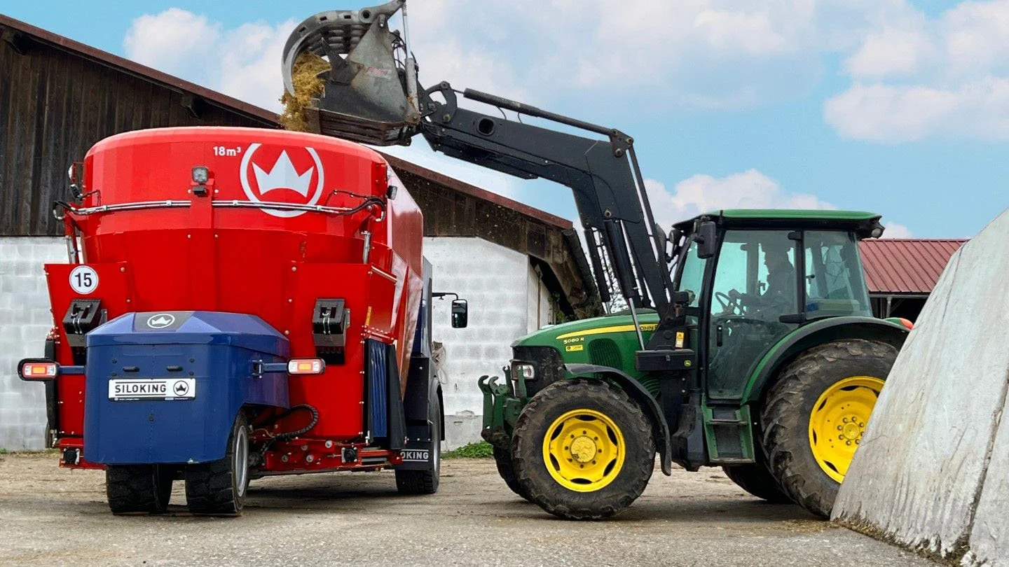 Rear view of an 18 cubic meter Siloking TruckLine eTruck 2012 being loaded with feed by a green John Deere tractor with a front loader bucket.