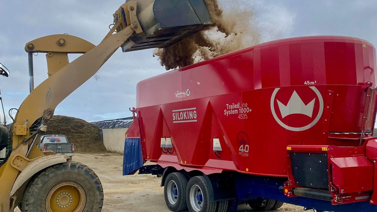 A heavy-duty yellow CAT wheel loader dumps a large bucket of feed into a massive 45 cubic meter red Siloking TrailedLine System 1000 plus 4535 triple-auger vertical mixer wagon at a farm silage bunker.