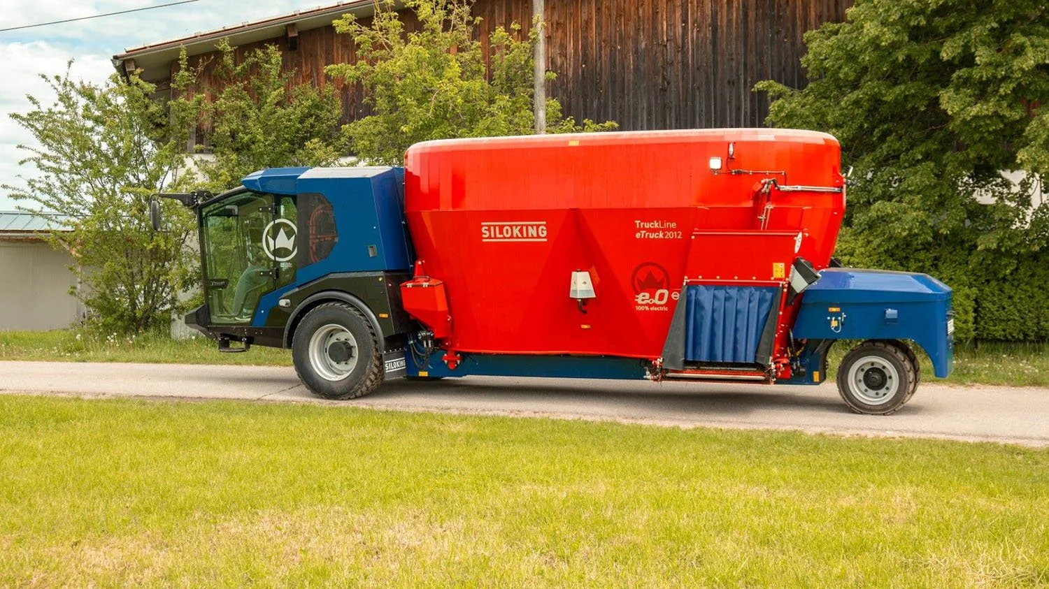 Left-side profile view of an all-electric red and blue Siloking TruckLine eTruck 2012 self-propelled mixer wagon traveling down an open farm lane.