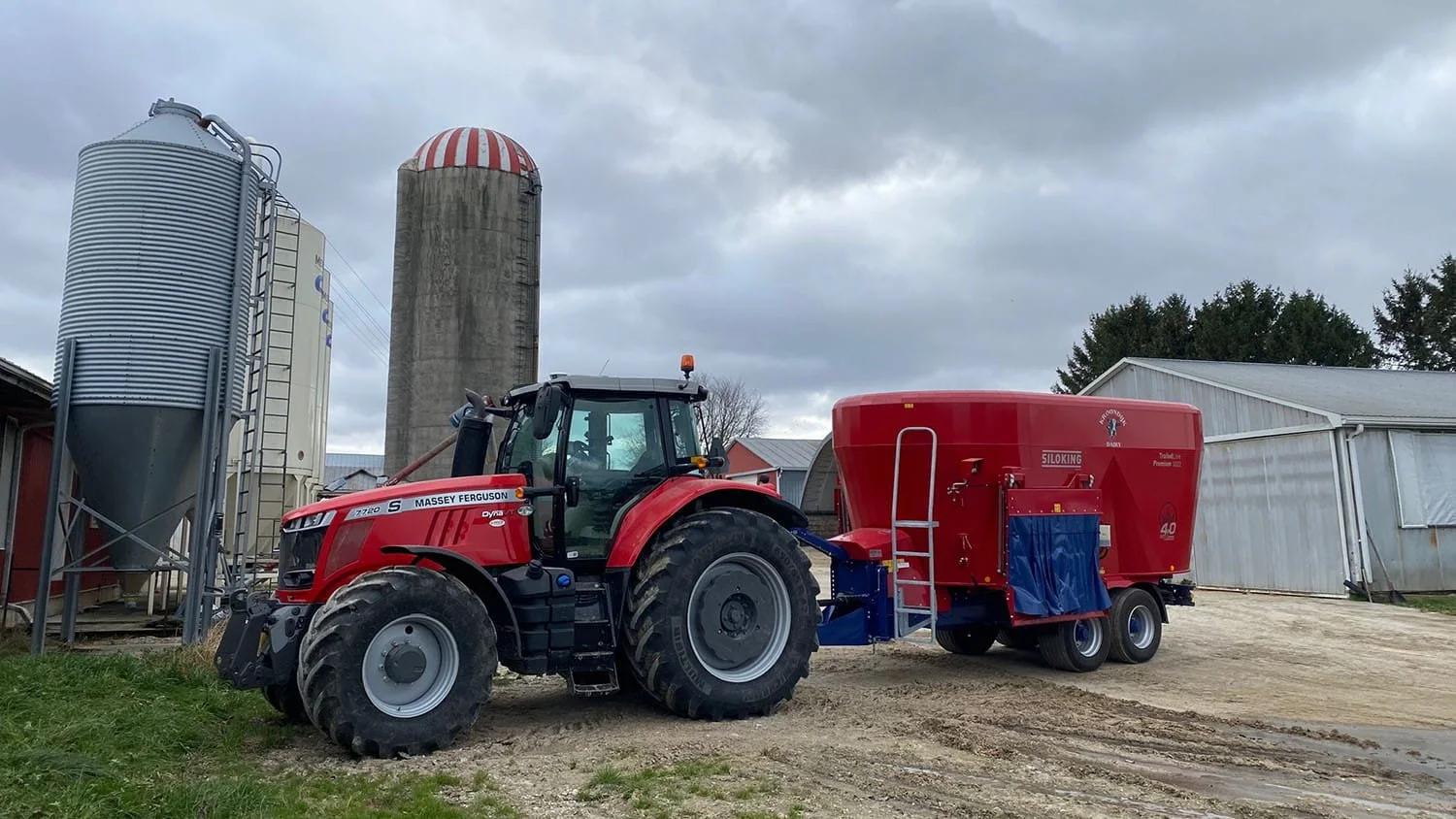 A large red Siloking TrailedLine Premium 3022 vertical feed mixer hitched to a red Massey Ferguson tractor parked next to concrete silos under an overcast sky.