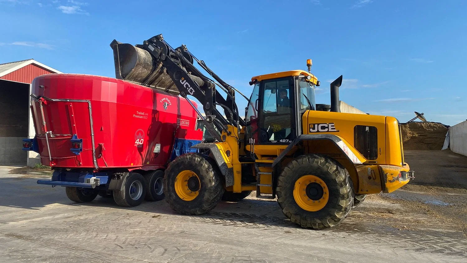 A yellow JCB wheel loader dumping feed ingredients into a massive twin-auger Siloking TrailedLine Premium 3022 vertical mixer wagon in a concrete bunker yard.