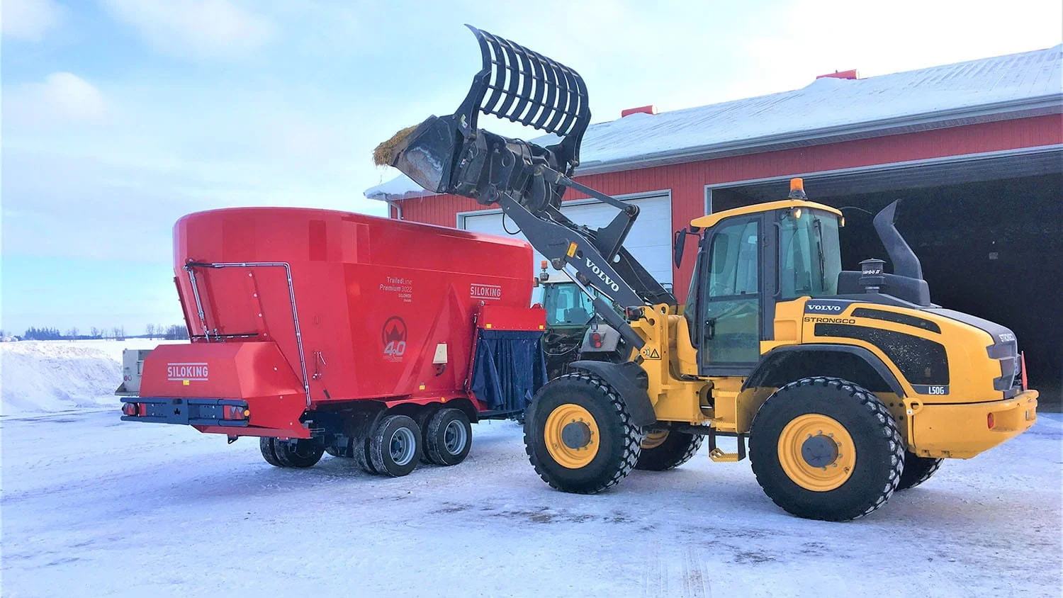 A yellow Volvo wheel loader with a silage bucket loading feed components into a twin-auger Siloking TrailedLine Premium 3022 vertical mixer wagon on a snow-covered Canadian farm lot in winter.
