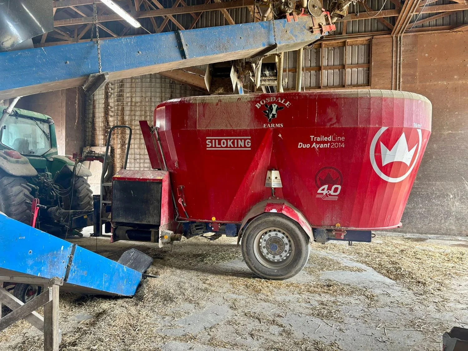 Side profile of a 16 m3 Siloking TrailedLine Duo Avant 2014 pull-type vertical TMR mixer hitched to a John Deere tractor inside a dairy barn.