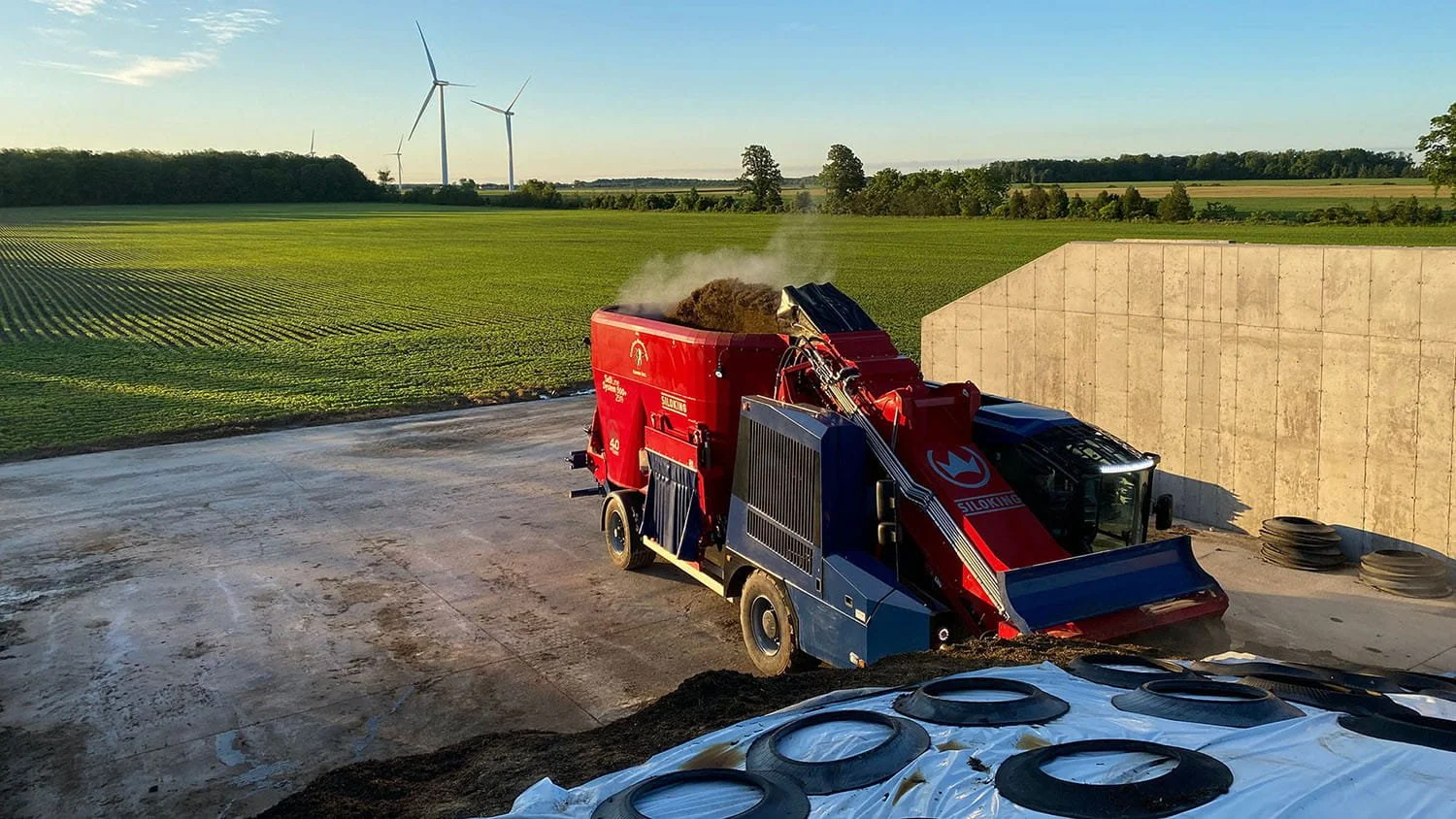 A high-angle view of a red and blue Siloking SelfLine System 500 plus 2519 self-propelled feed mixer demonstrating bunker silage extraction with its milling arm alongside an open crop field with wind turbines.