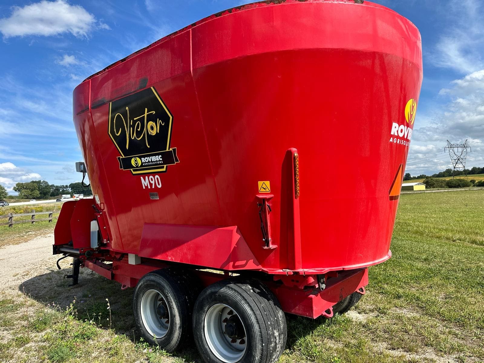 Rear three-quarter view of a red Rovibec Victor M90 twin auger vertical mixer wagon on a tandem axle in a grass field.