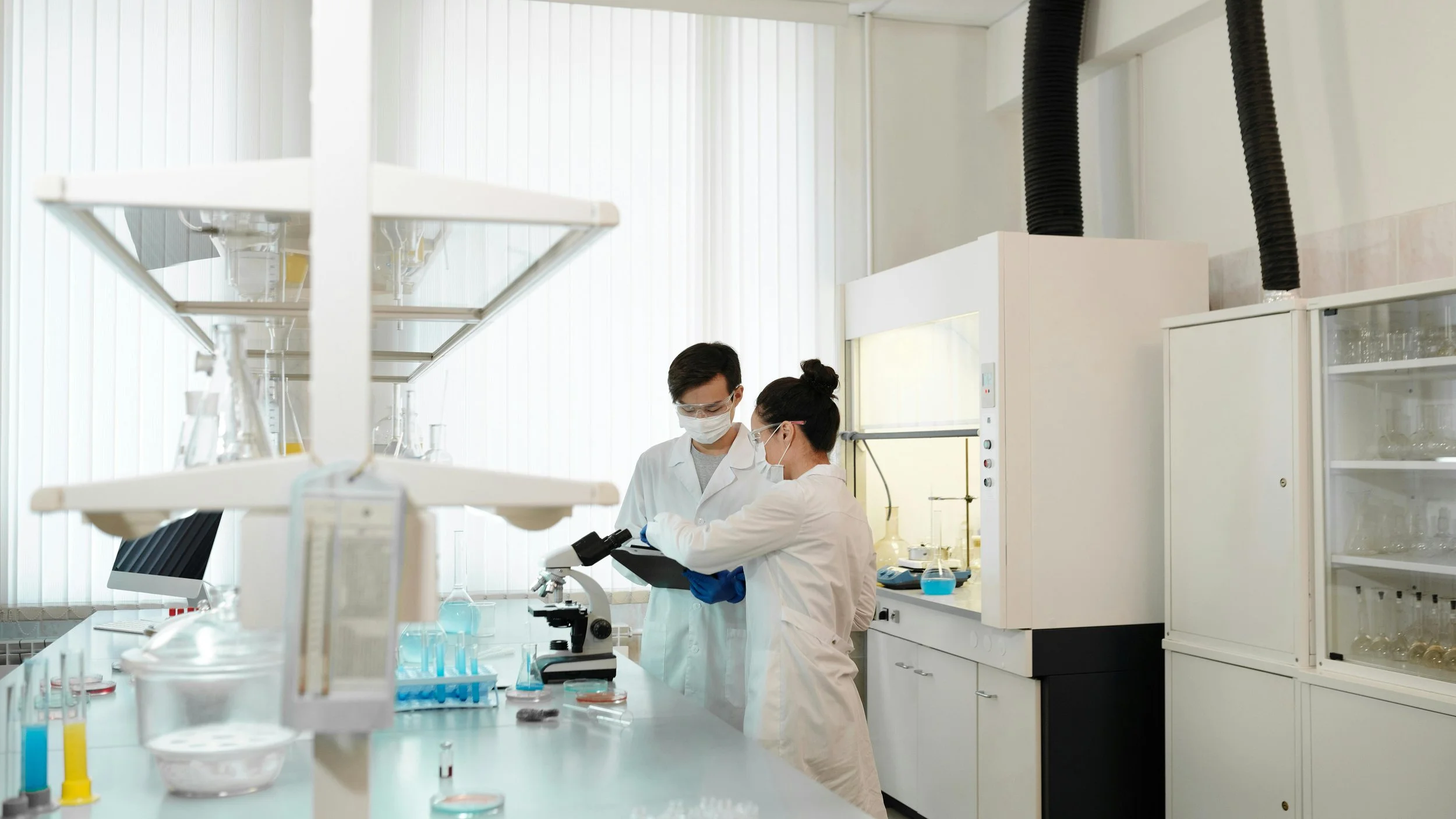Two scientists wearing white coats, masks, and goggles are working in a laboratory with scientific equipment, including a microscope, test tubes, and chemicals.