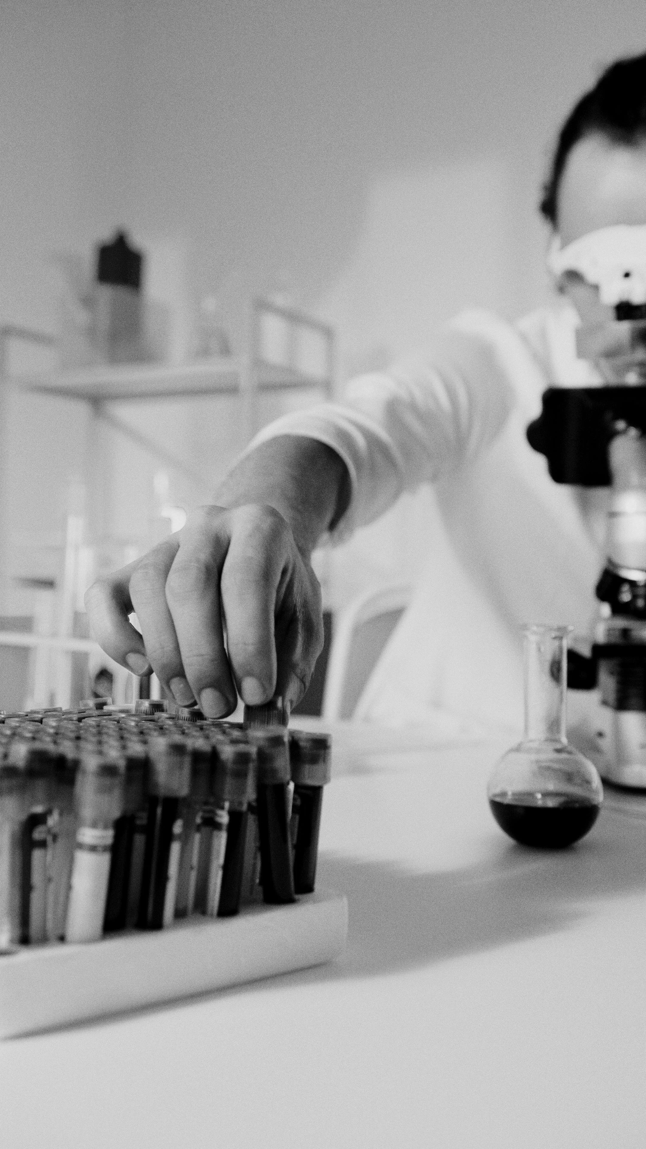 A scientist in a laboratory working with test tubes and a microscope, using a pipette, with a beaker of dark liquid nearby.