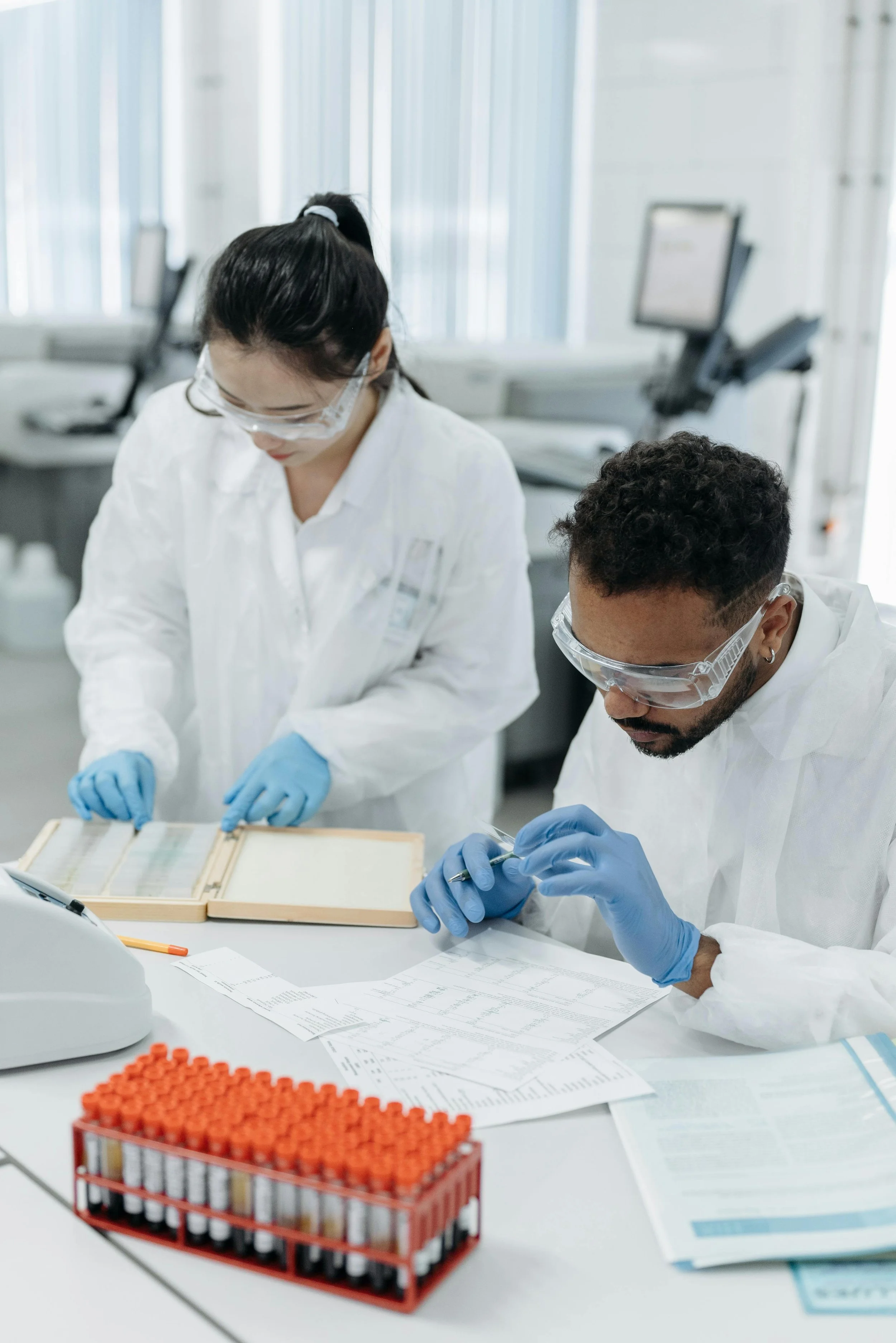 Two scientists wearing protective lab gear working at a lab bench with test tubes, papers, and lab equipment.