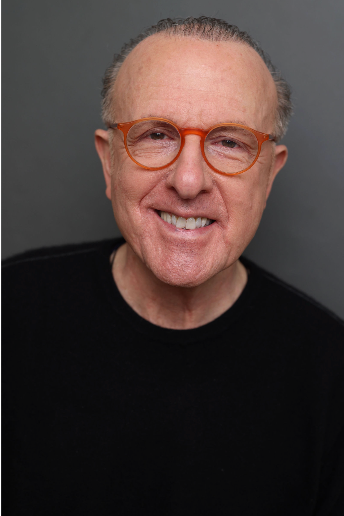A smiling middle-aged man with short gray hair, wearing orange glasses and a black shirt, standing against a gray background.