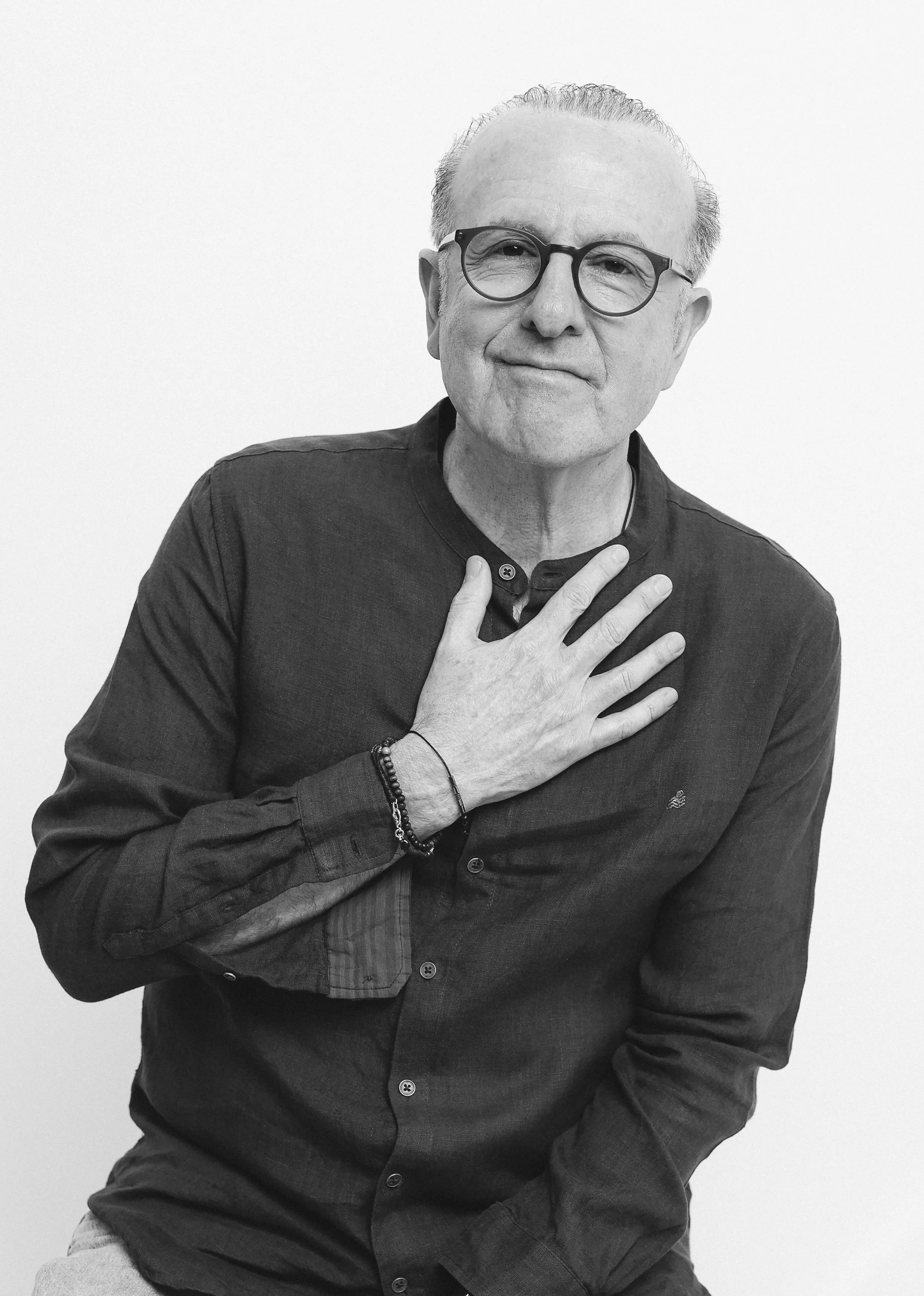 A man with glasses and grey hair, wearing a dark long-sleeve shirt, holds one hand on his chest and looks at the camera with a gentle expression against a plain background.