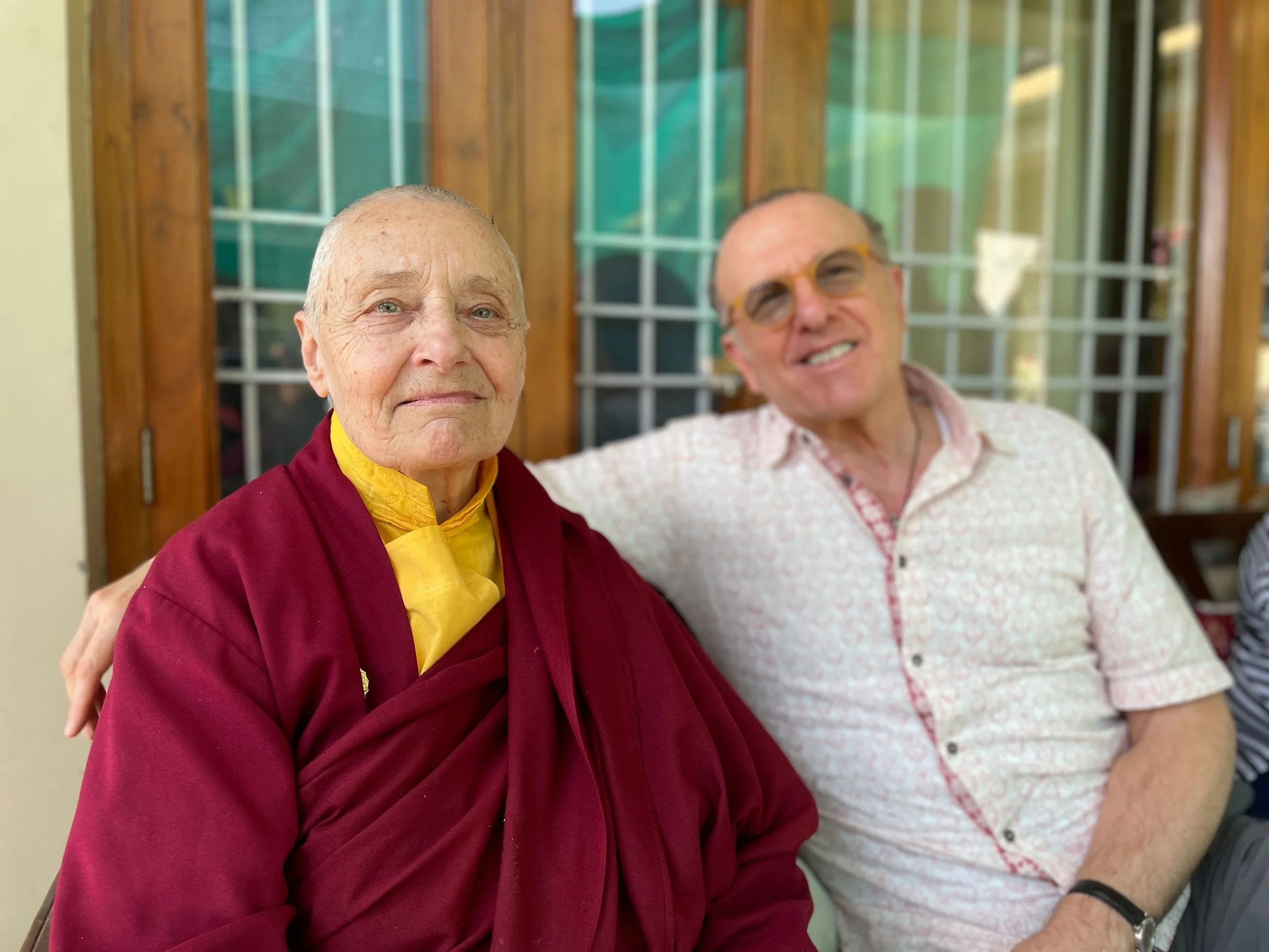 A Buddhist nun in maroon robes sitting next to a man in a short-sleeved, button-up shirt, both smiling, with a wooden-framed glass door in the background.