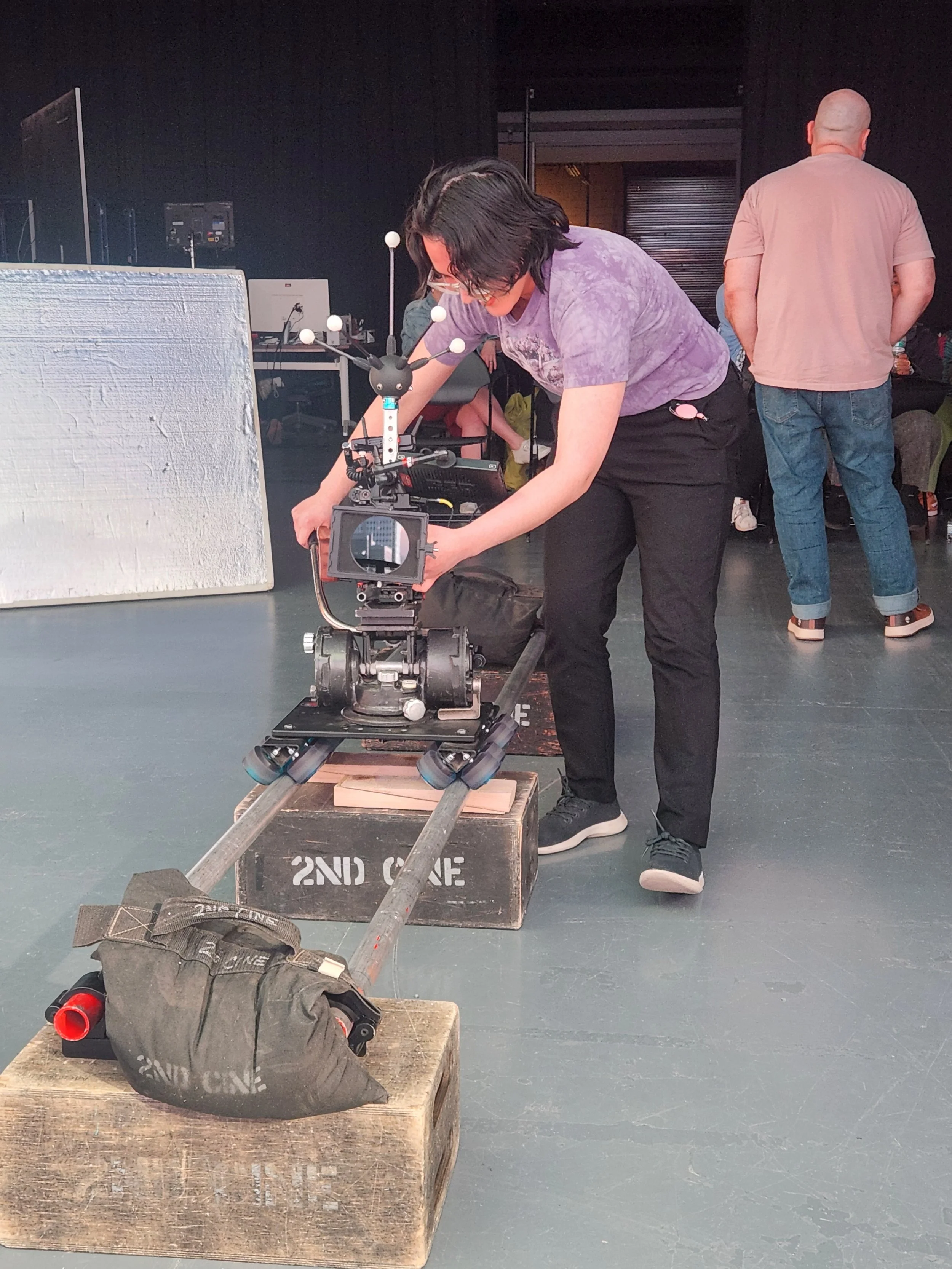 A person with dark hair and glasses is operating a camera mounted on a dolly, set on a wooden platform. In the background, other people are seated or standing in a dark room, with some equipment and props around.