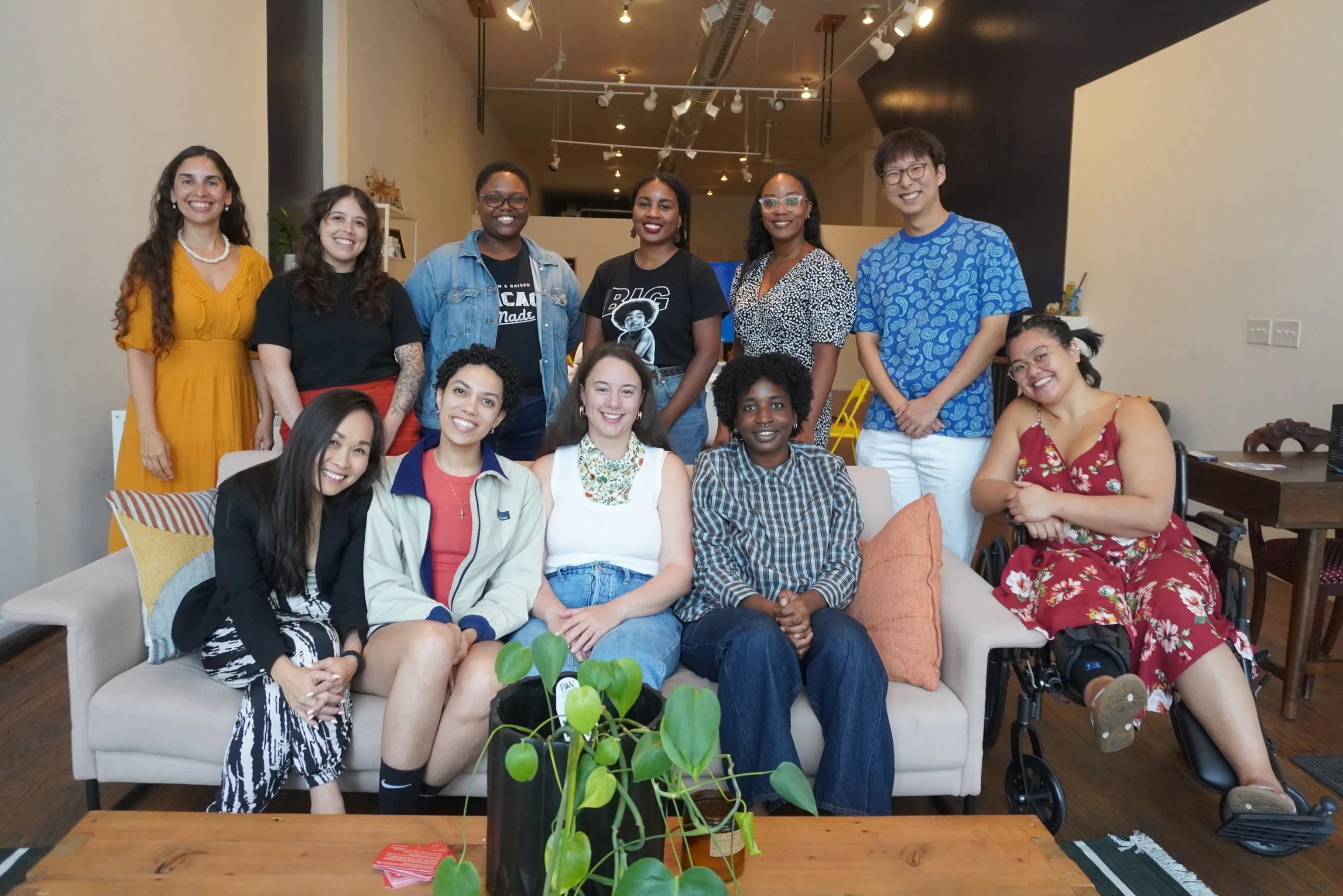 A group of twelve gathered together indoors, smiling for a photo. The group is seated and standing on a sofa and chairs, with a plant and some decorative pillows in the foreground. The background features a beige wall, some bookshelves, and lighting fixtures.