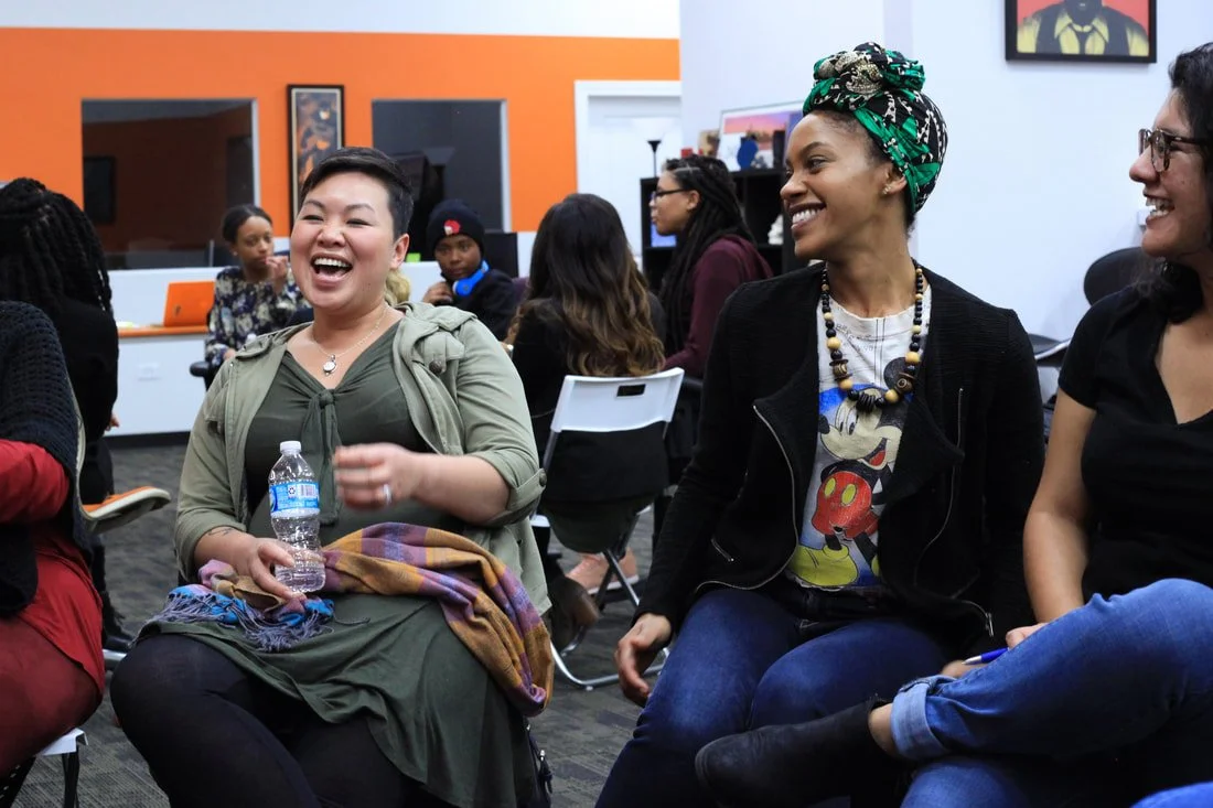 Group of women in a room, smiling and laughing, seated on white chairs, with a colorful background and some artwork on the wall.