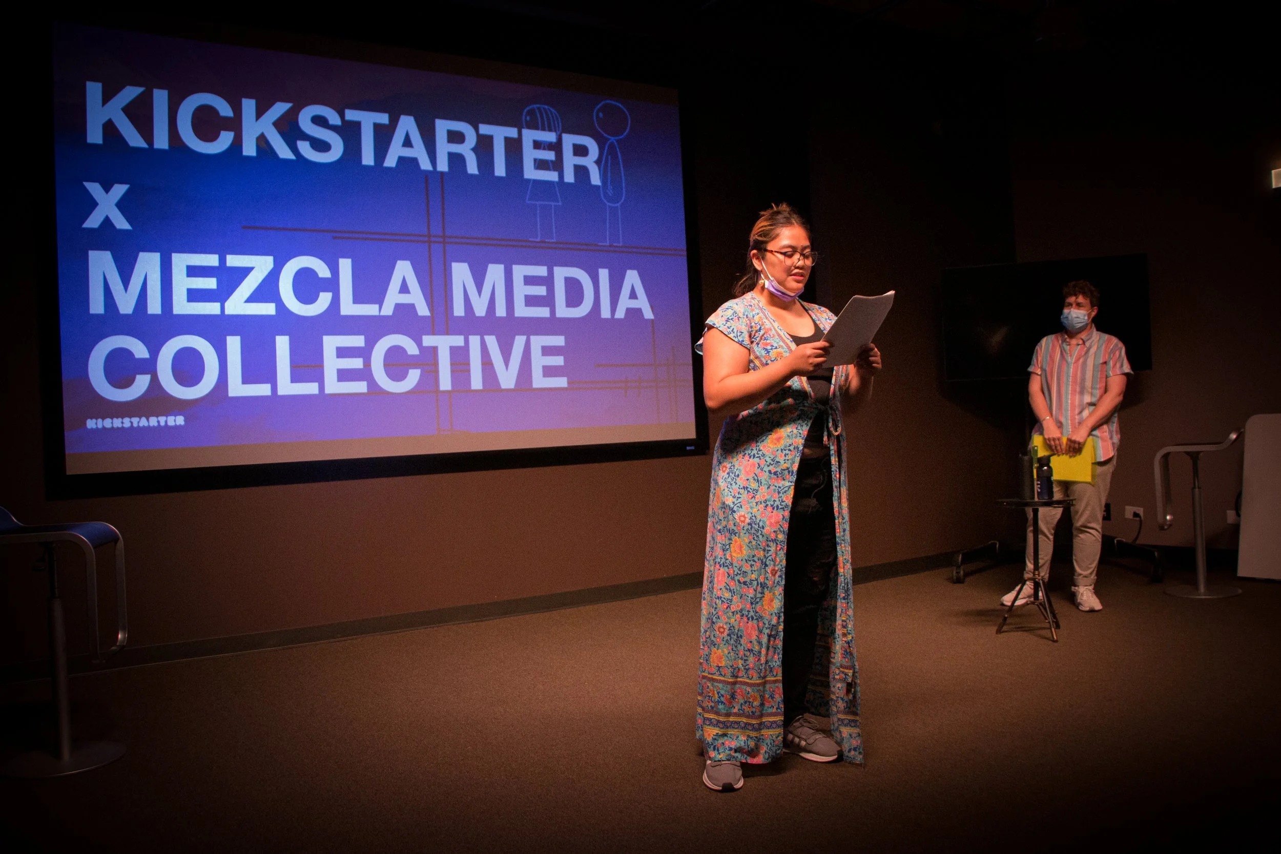 A person in a colorful floral dress and glasses standing on stage reading from a paper, with a man in striped shirt and mask standing behind them, in front of a large screen displaying 'Kickstarter x Mezcla Collective' logo.