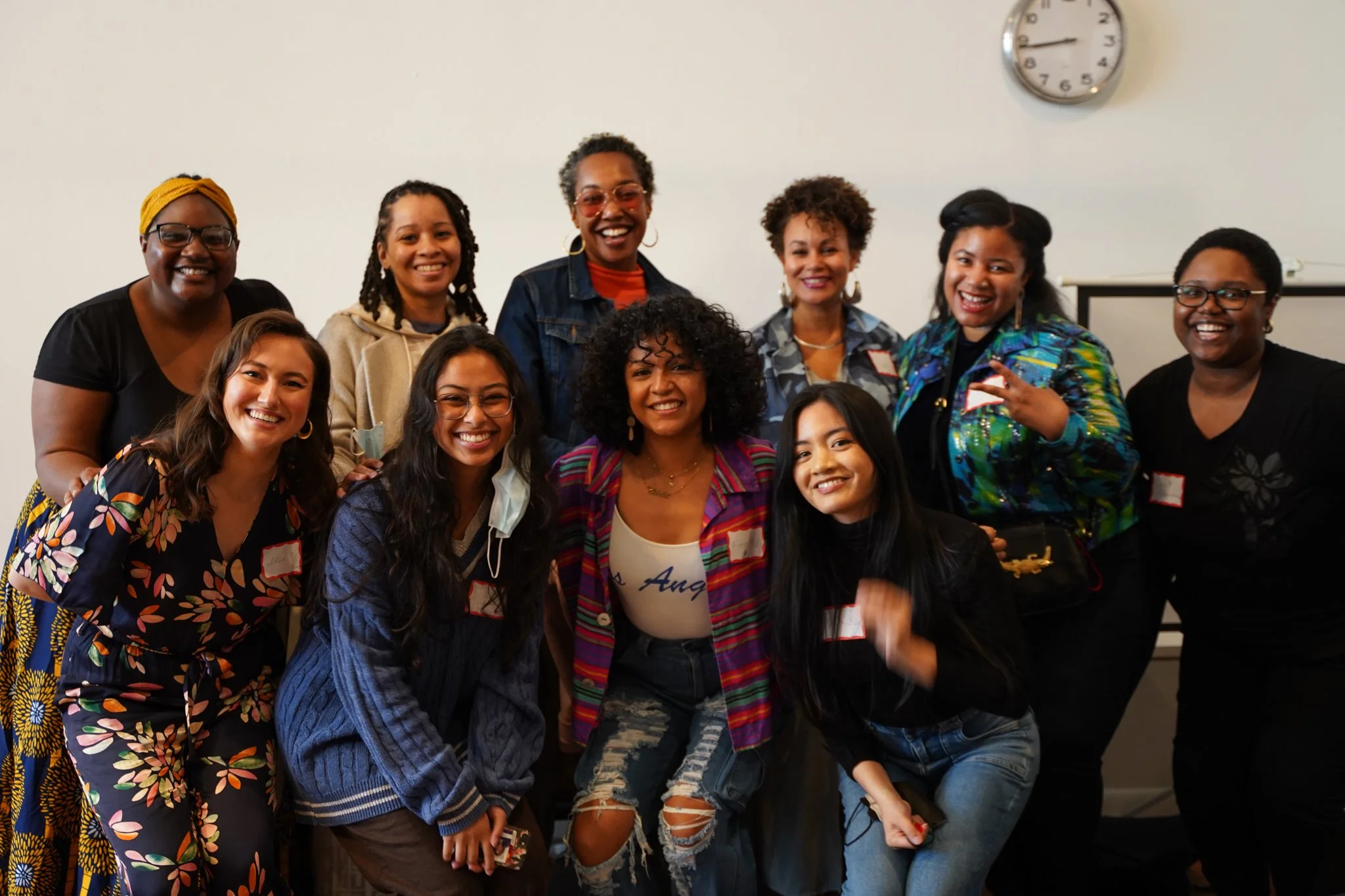 A group of ten smiling people pose for a photo indoors, some wearing name tags, with a wall clock showing 3:14 in the background.