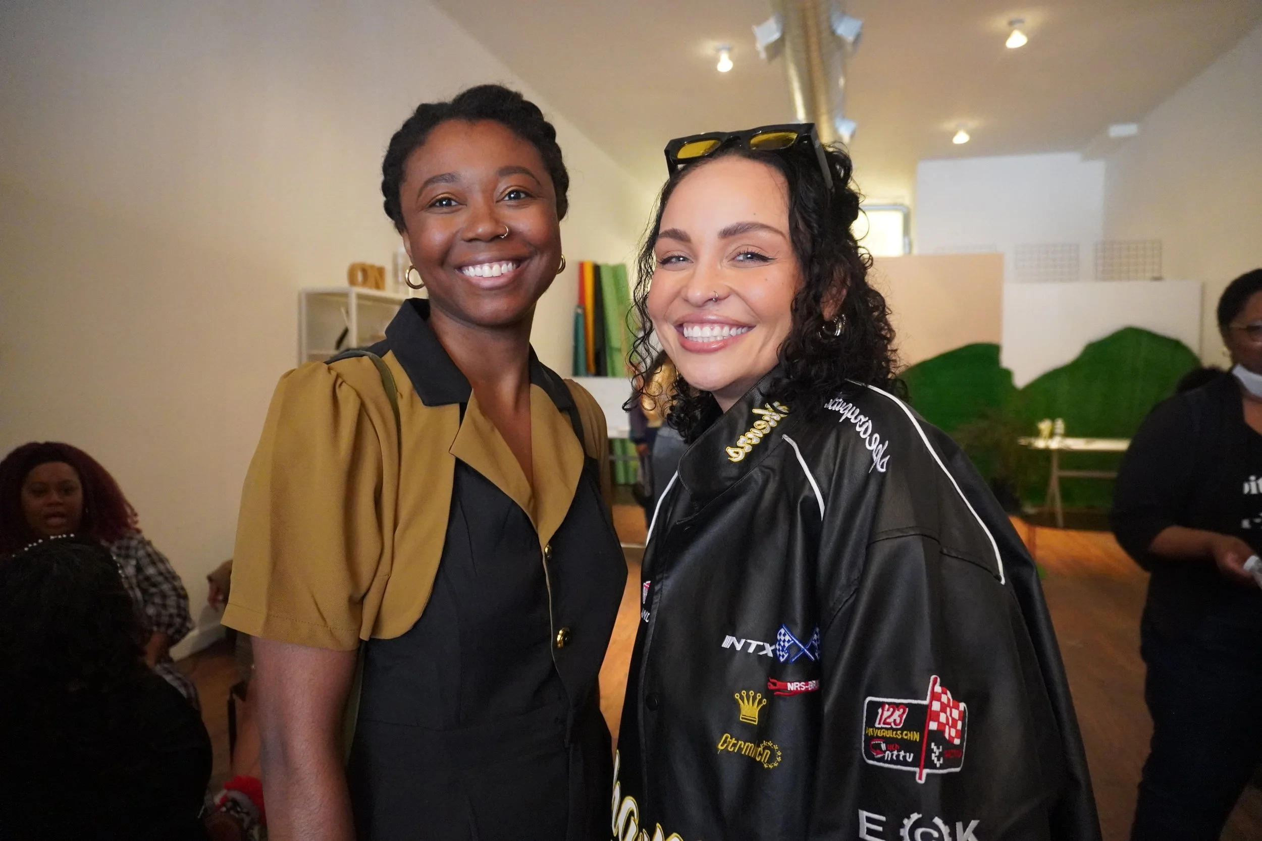 Two women smiling, one with dark skin and short braided hair wearing a tan jacket over black, the other with light skin, curly black hair, and sunglasses on her head, wearing a racing jacket. They are indoors with other people and colorful books in the background.