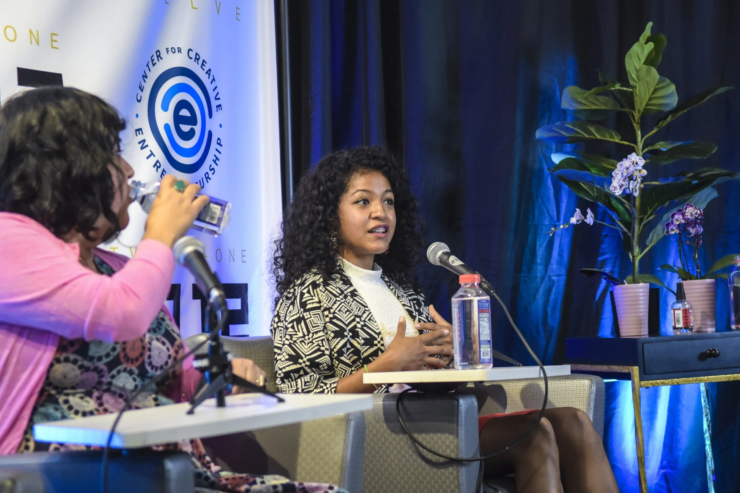 A woman with curly black hair speaking into a microphone at a panel discussion. She's wearing a patterned black and white blazer and a cream sweater. To her left, a woman in a pink jacket with glasses is drinking water. The panel is seated at a table with microphones and water bottles. Behind them is a large banner with a blue logo and text, and there are potted plants and dark curtains in the background.