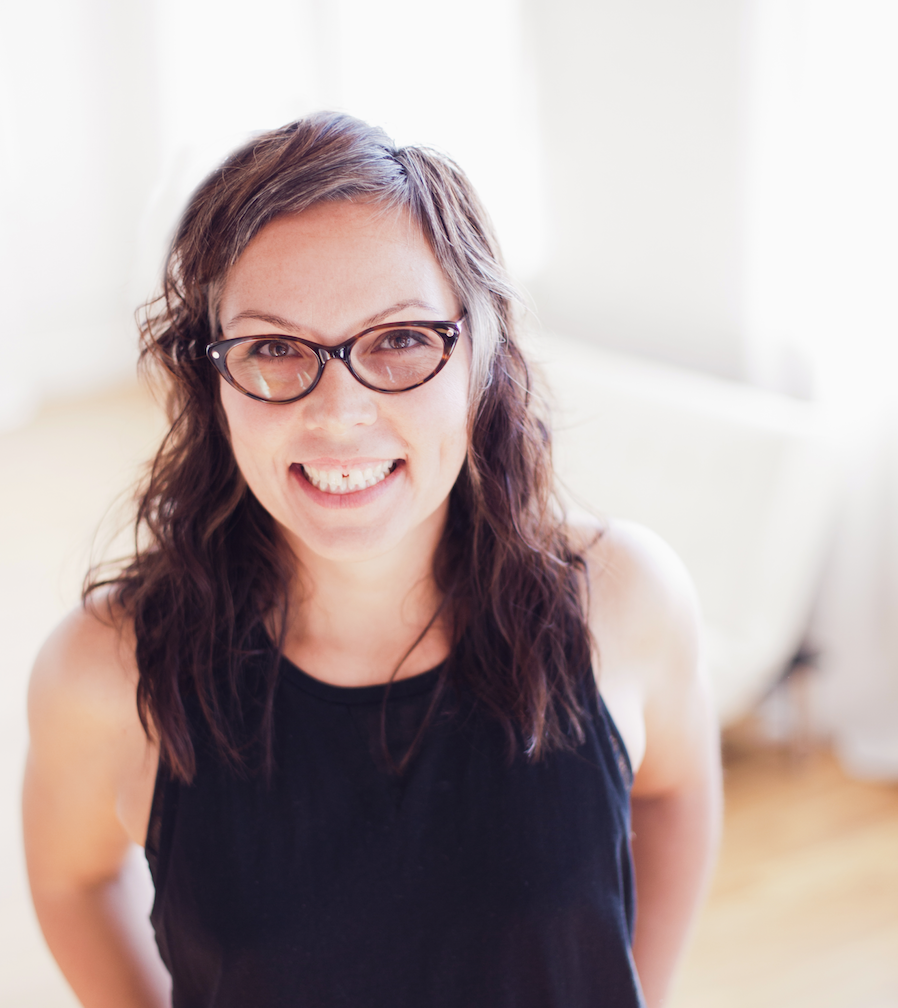 A woman with glasses and long, wavy hair smiling at the camera, wearing a black sleeveless top, in a bright room.