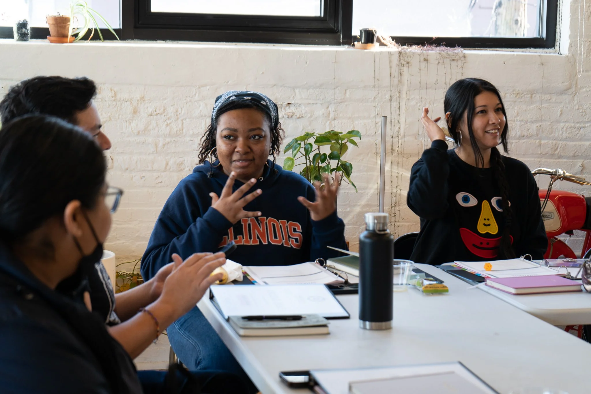 A group of four people sitting at a table in a bright room with a large window. They are engaged in conversation with notebooks, papers, and drinks on the table.