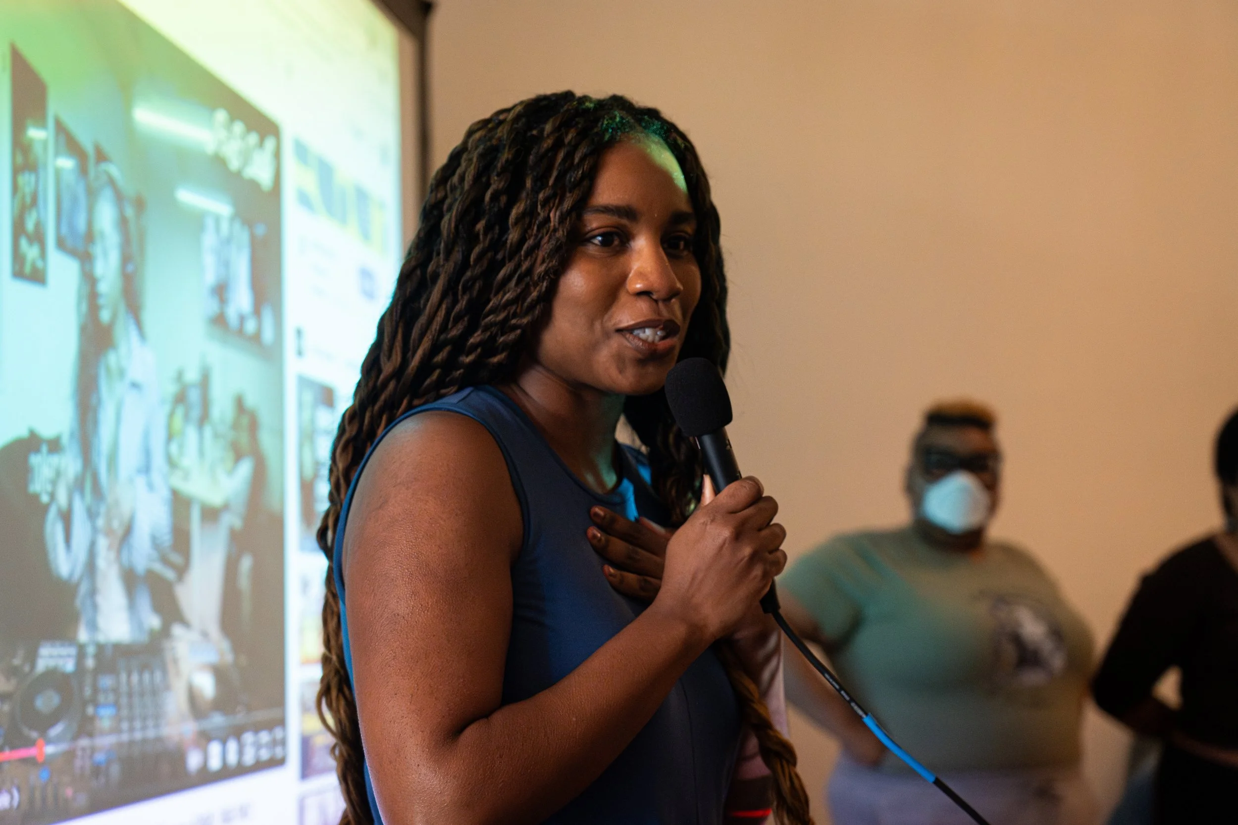 A woman with long braided hair is speaking into a microphone in front of a projection screen, with two people wearing masks in the background.